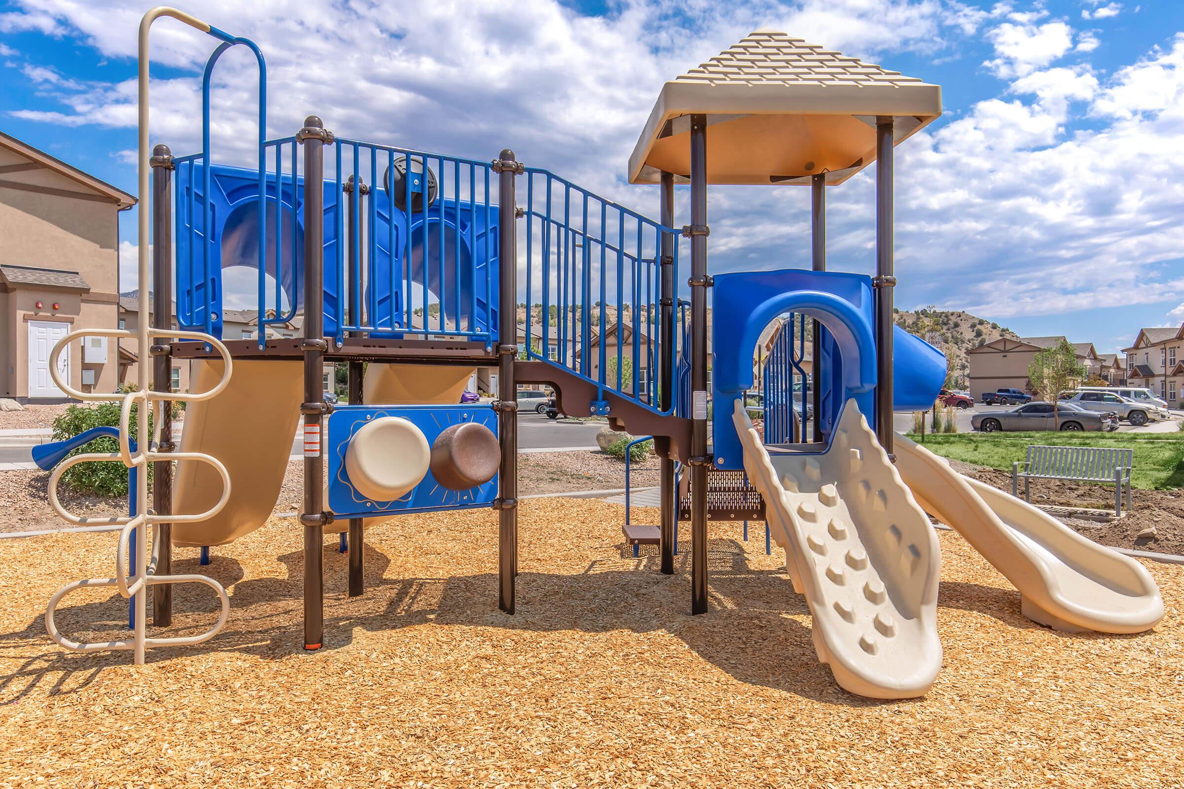 A colorful playground structure featuring two slides, climbing elements, and interactive panels, surrounded by a sandy area. The background shows residential buildings and a clear blue sky with scattered clouds.