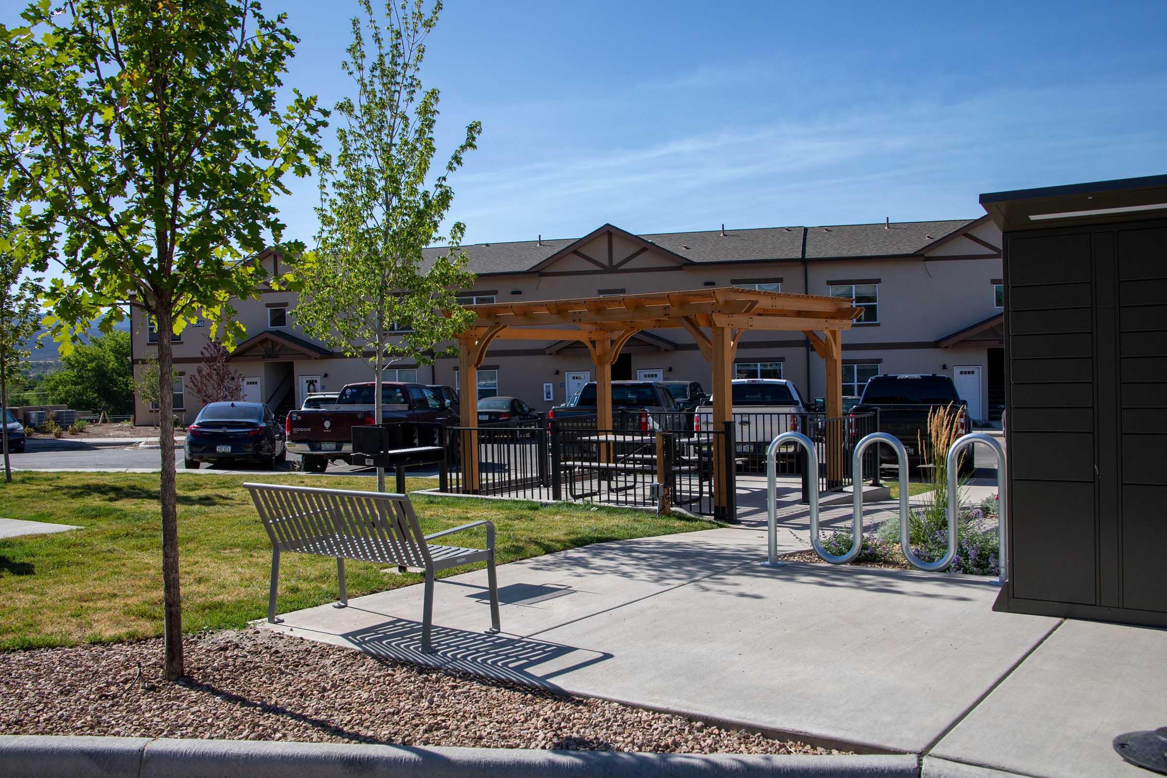 A sunny outdoor area featuring a wooden gazebo, benches, and bike racks. Surrounded by grass and trees, there are parked cars in the background, and multi-story buildings with a clear blue sky overhead. The setting offers a communal space for relaxation and recreation.