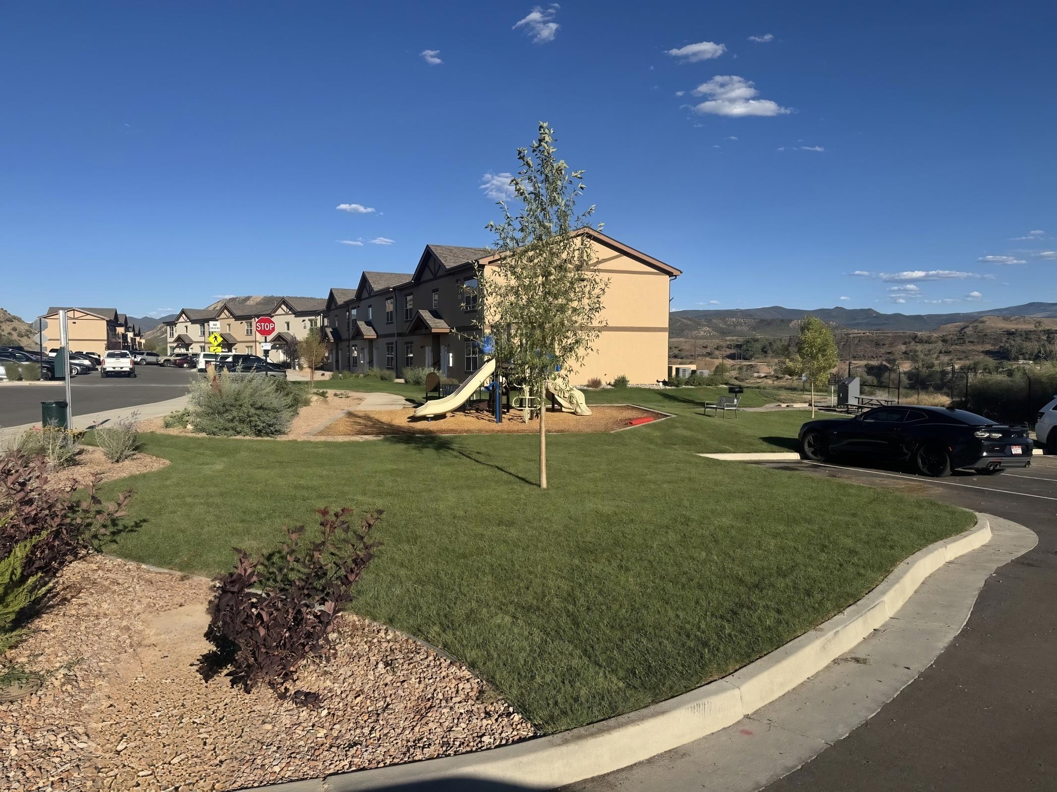 A residential area featuring a grassy lawn, a playground with a slide, and several multi-unit buildings. In the background, there are mountains and a clear blue sky. A few cars are parked nearby, and the landscape includes shrubs and decorative rocks.