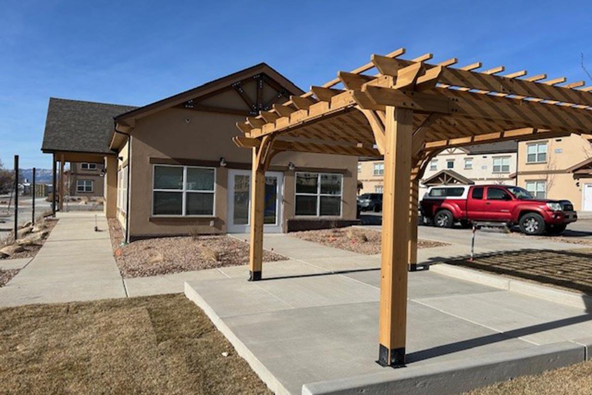 A building with a light brown exterior and multiple windows, featuring a wooden pergola in the foreground. The landscape includes a paved pathway and some grass. A red pickup truck is parked nearby, and a row of similar buildings is visible in the background under a clear blue sky.