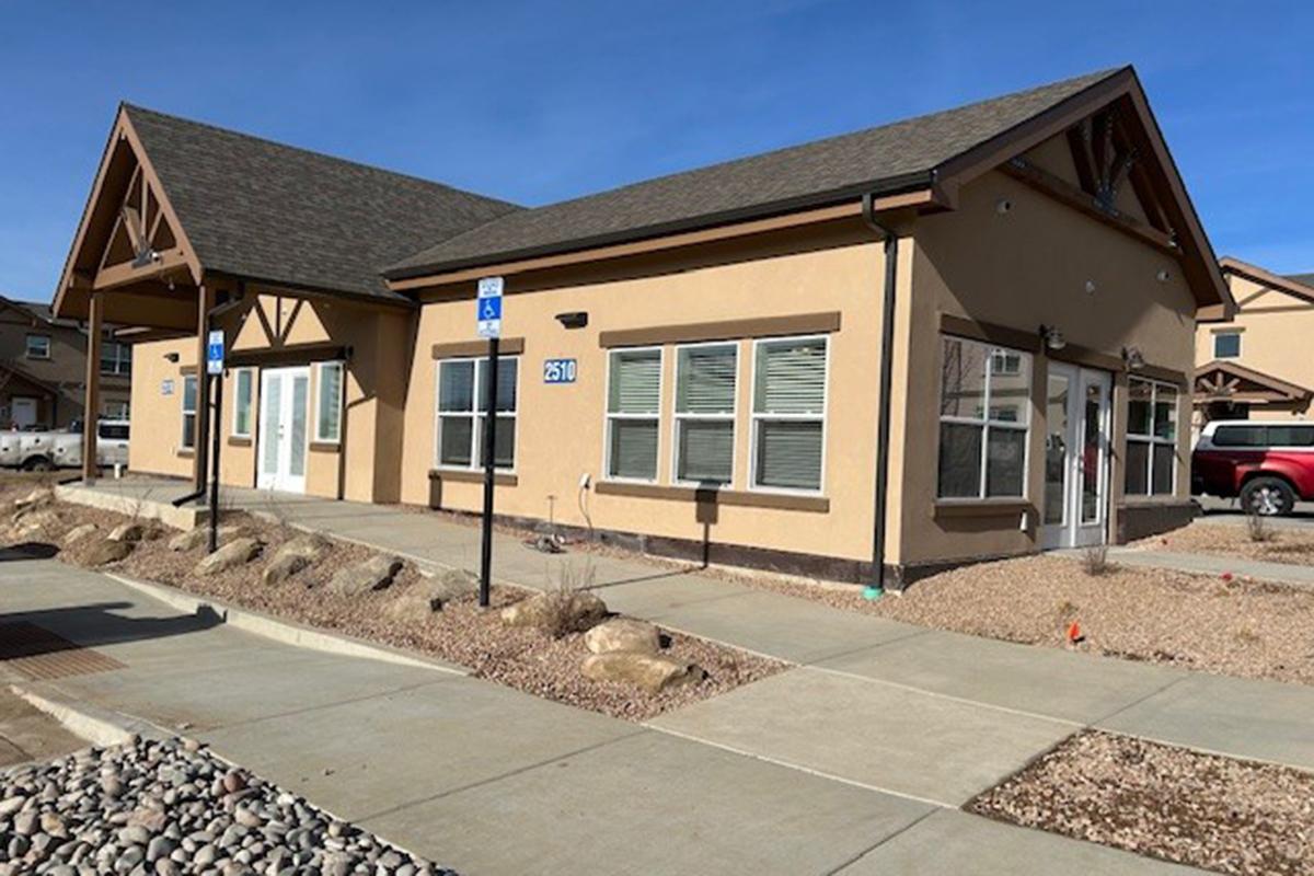 A single-story building with a tan exterior and a brown roof, featuring large windows, a covered entrance, and accessible parking spaces in front. Surrounding the building is a paved walkway and landscaped areas with gravel and low shrubs, under a clear blue sky.