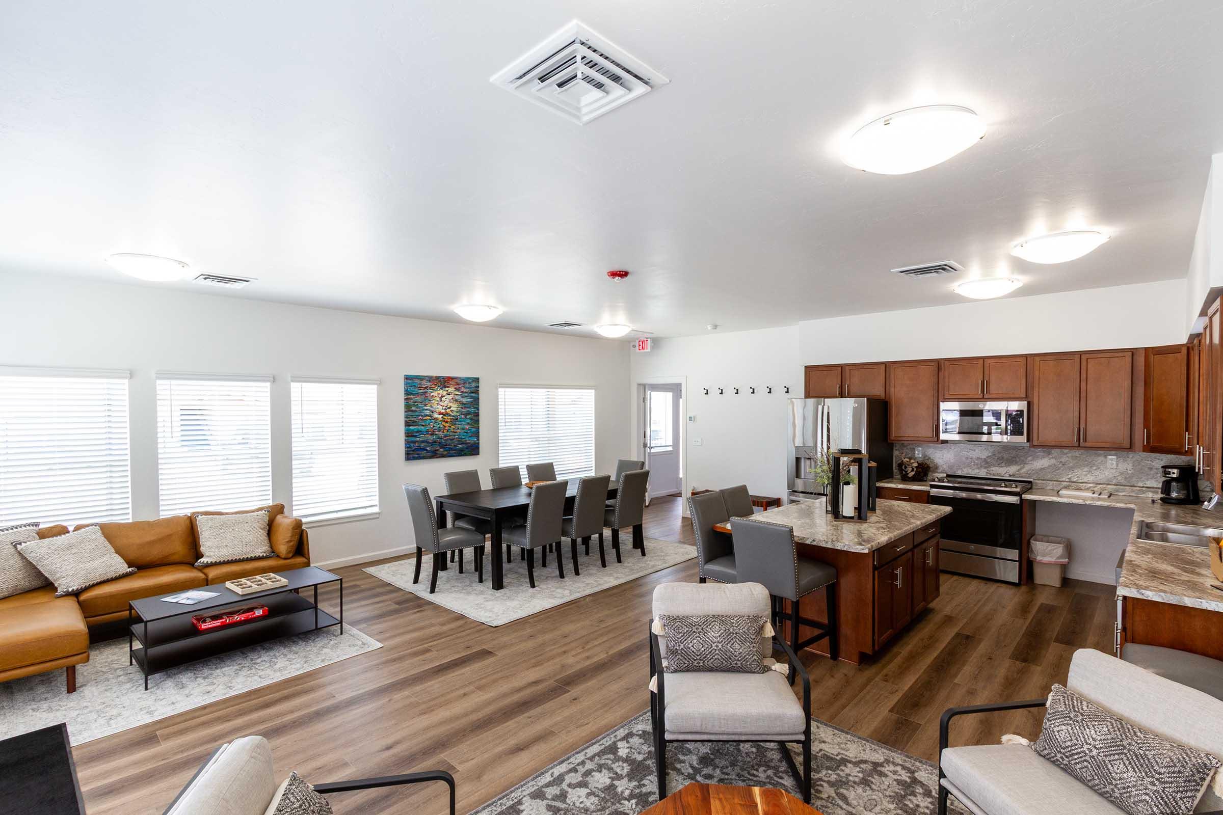 A spacious and modern common area featuring a brown leather sofa, a dining table with chairs, and a kitchen with wooden cabinets. Large windows let in natural light, and the flooring is a combination of wood and carpet. Decorative artwork adorns one wall, adding a vibrant touch to the space.