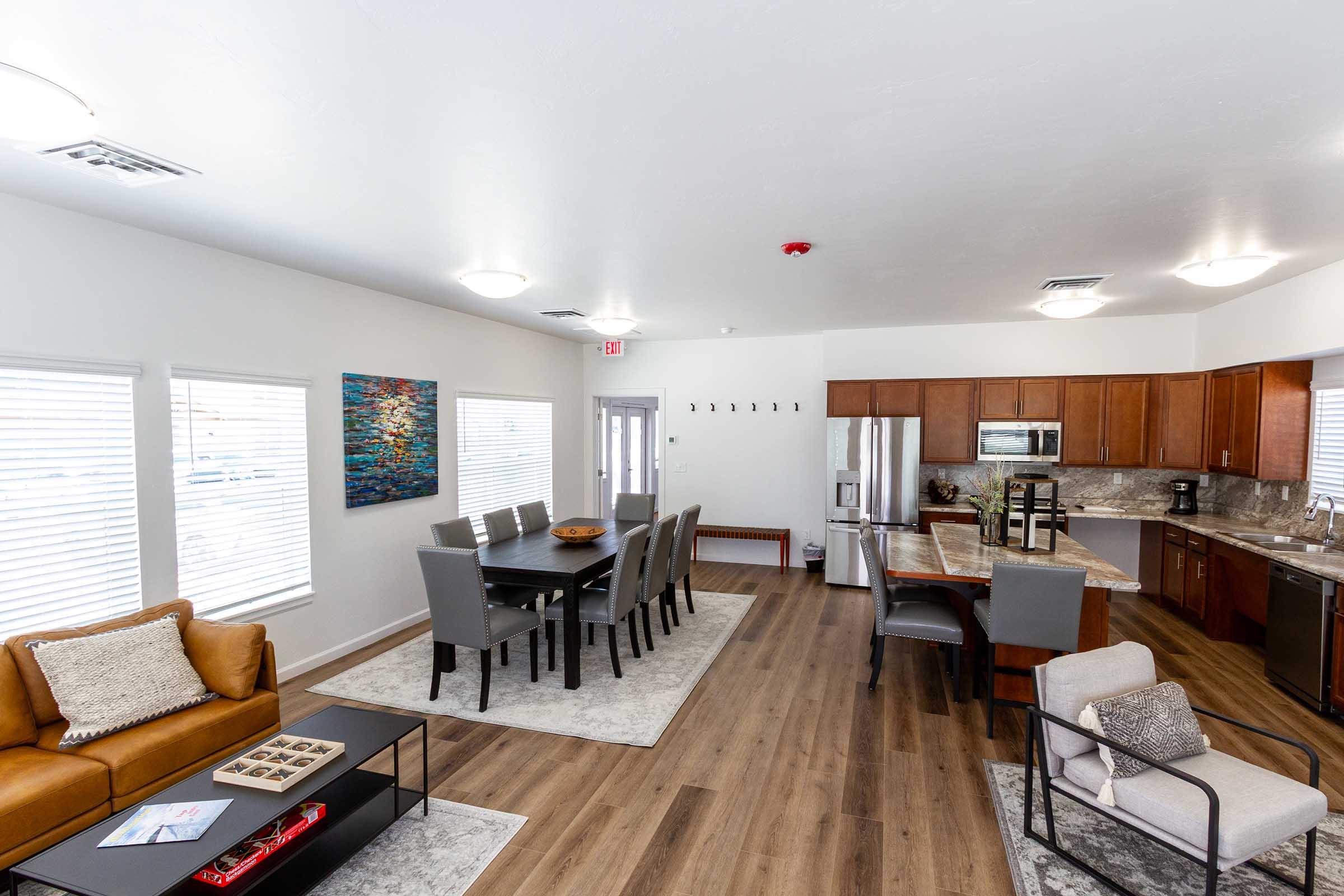 A modern, spacious living area featuring a brown leather sofa, a gray armchair, and a dining table with gray chairs. The kitchen area includes stainless steel appliances and wooden cabinetry. Large windows provide natural light, and artwork decorates the wall, adding color to the room.