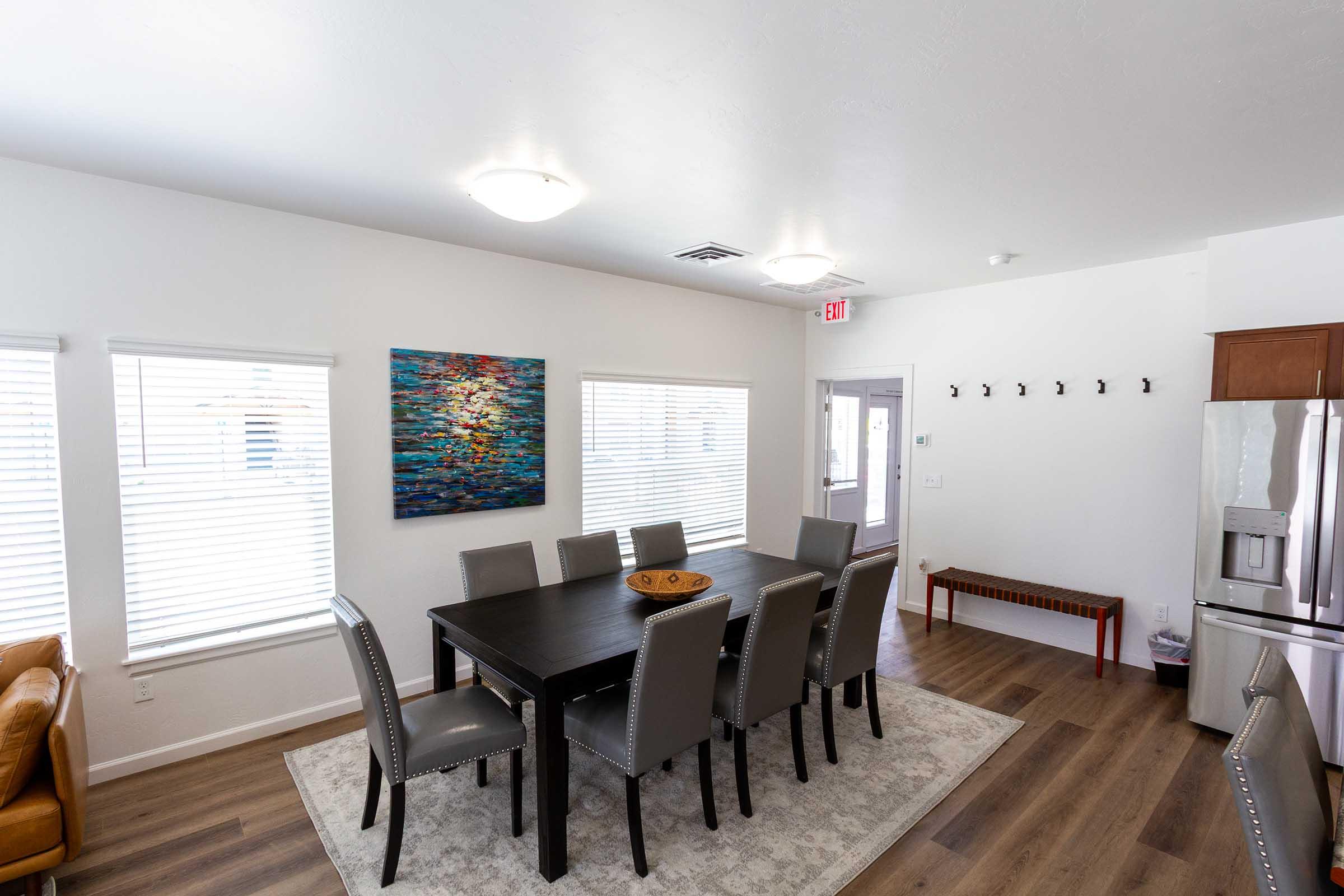 A modern dining area with a large black table surrounded by gray upholstered chairs. A colorful abstract painting hangs on the wall, complemented by large windows with blinds. A small wooden bench and hooks line one wall, with a kitchen visible in the background. The space is bright and inviting.
