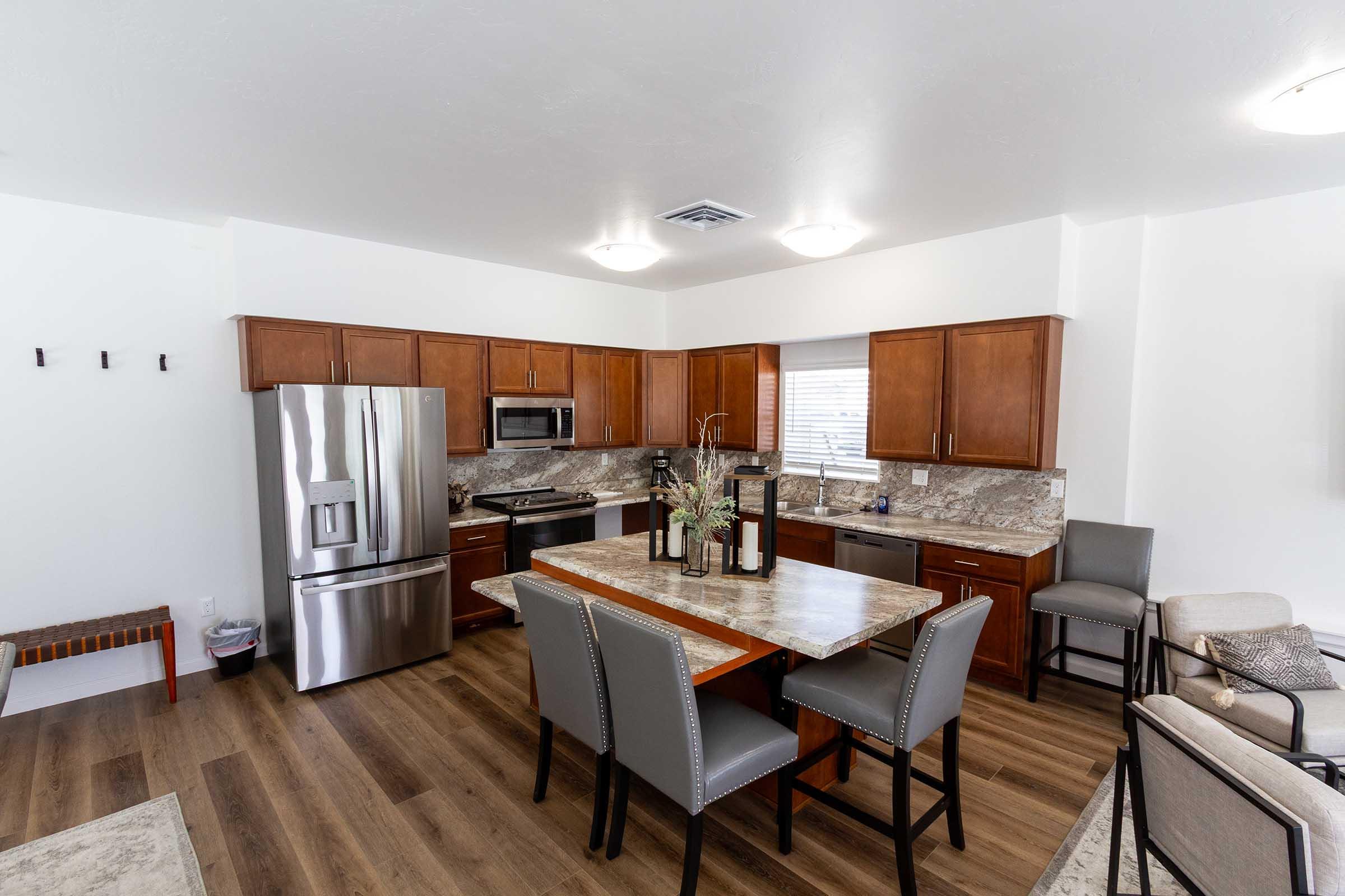 Modern kitchen featuring wooden cabinetry, stainless steel appliances, and a large granite island with seating. Light-colored walls and hardwood flooring create a bright, inviting atmosphere. The space includes a dining area with decorative items on the table and a cozy seating area in the background.