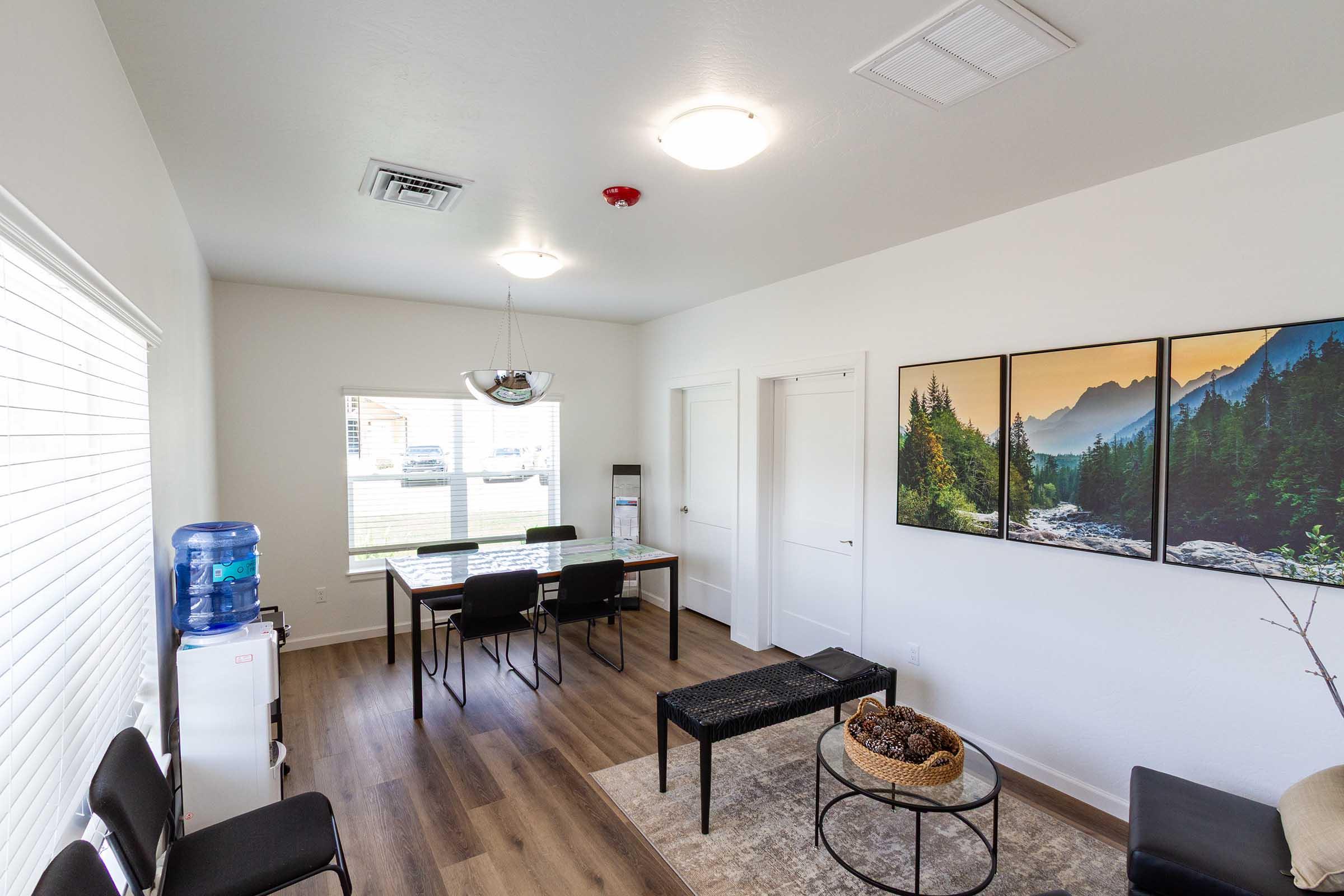 A modern office waiting area featuring a simple design with a table and chairs, a water cooler, and two framed landscape photographs on the wall. The space has natural light from a window, light-colored walls, wood flooring, and a decorative bowl on a bench.