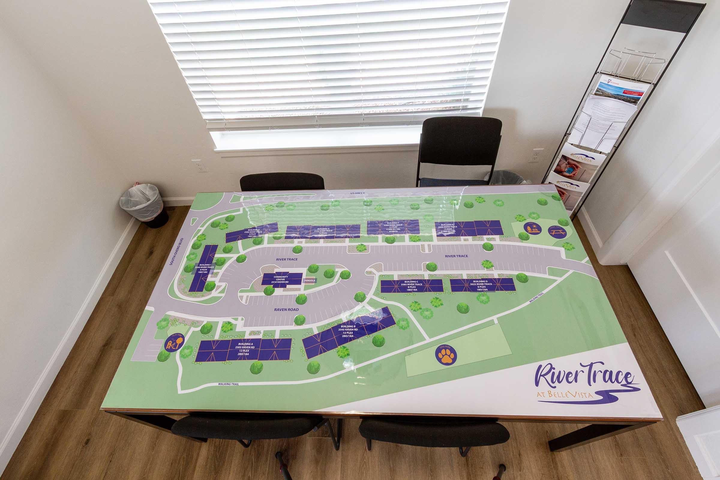 A top-down view of a large table featuring a detailed layout map of River Trace at Sunny Vista, including buildings, parking areas, and landscaping. The table is positioned in a bright room with a chair and a brochure display in the background.
