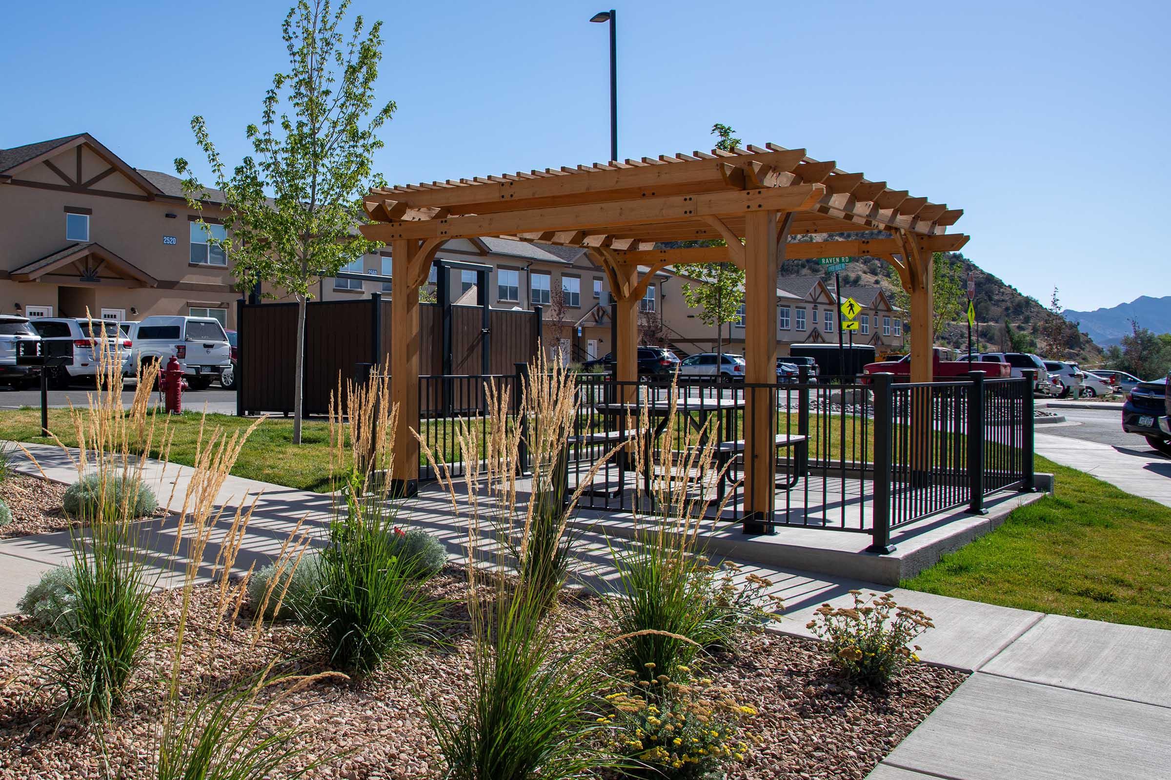 A wooden pergola with a picnic table underneath is set in a landscaped area featuring grasses and shrubs. In the background, there are residential buildings and parked cars. The sky is clear and blue, and there are trees around the structure.