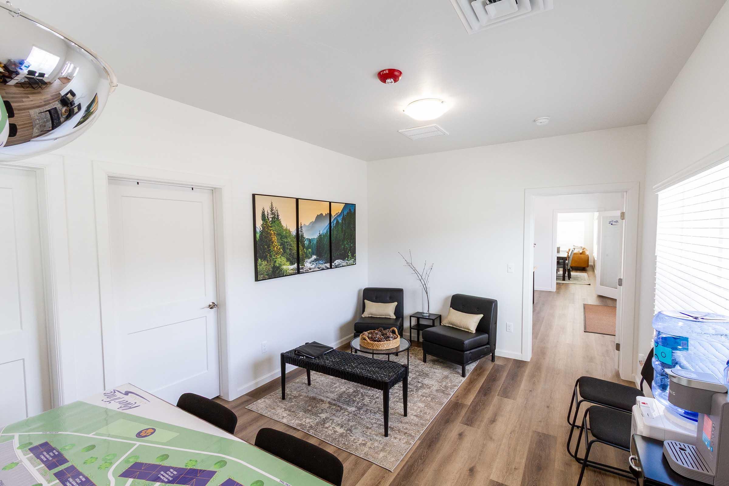 A well-lit interior showcasing a cozy living area with two black chairs, a small table with decorative items, and a framed mountain landscape on the wall. A table with a map is visible in the foreground, while a doorway leads to another room in the background.