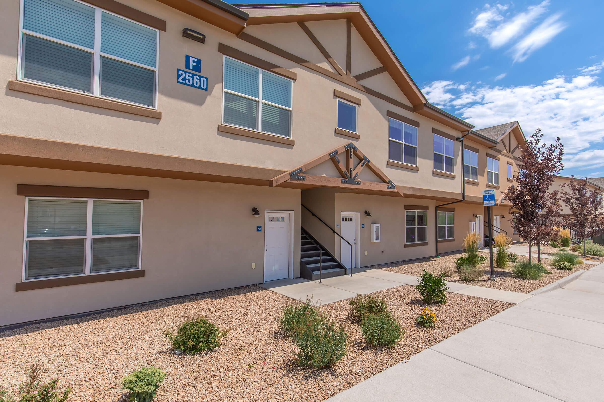 A modern two-story apartment building with light brown exterior, featuring multiple windows and doorways. The building has a clearly marked entrance with a staircase, surrounded by landscaped areas with small shrubs and gravel. Bright blue sky and scattered clouds are visible in the background.