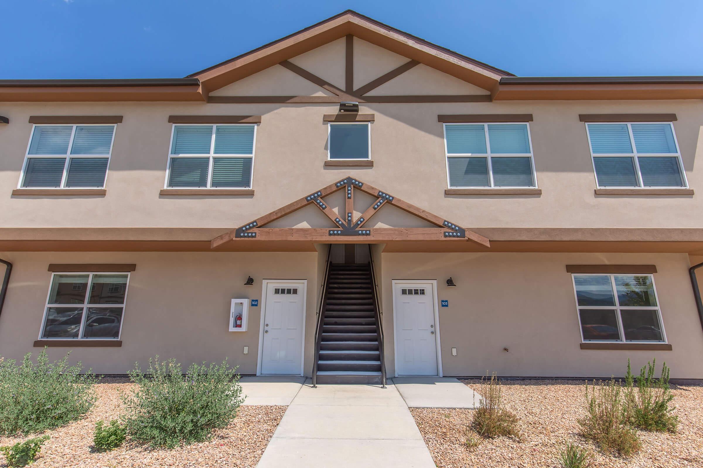 Two-story residential building with a staircase leading to the second floor. The exterior features beige walls, large windows, and decorative wooden beams. There are two white doors at the base, and the surrounding area has low shrubs and gravel landscaping. The sky is clear with minimal cloud cover.