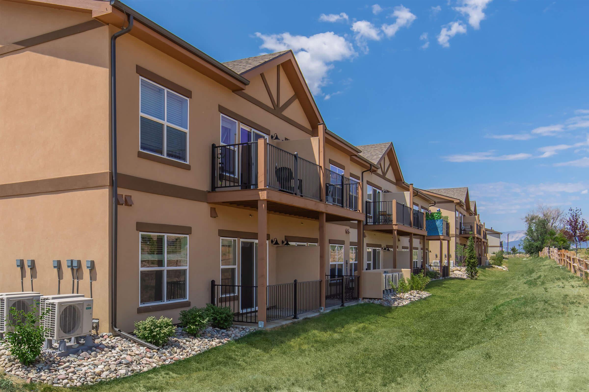 A row of modern townhouses with balconies, set against a bright blue sky. The buildings feature a combination of tan and brown exteriors, with neatly manicured grass and a gravel path. Air conditioning units are visible on the side, and a wooden fence lines the background.