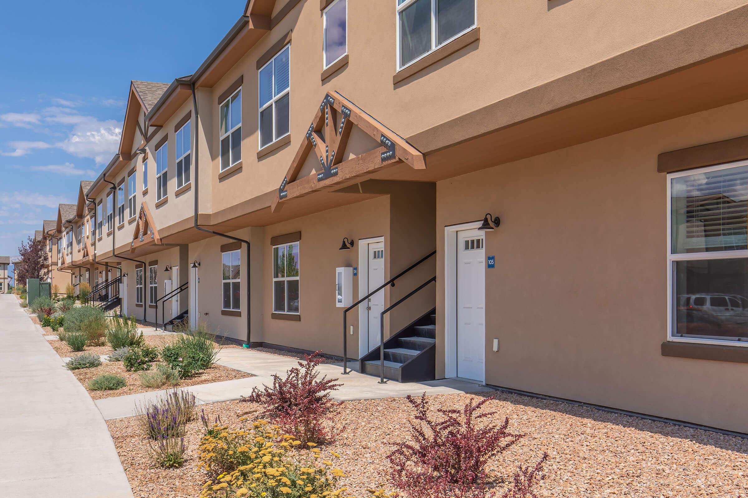 A row of modern townhouses with beige exteriors, decorative wooden accents, and black railings. Each unit features a front door with a small porch. The pathway in front is lined with low shrubs and flowers, leading to the main entrance. Bright blue sky with some clouds in the background.