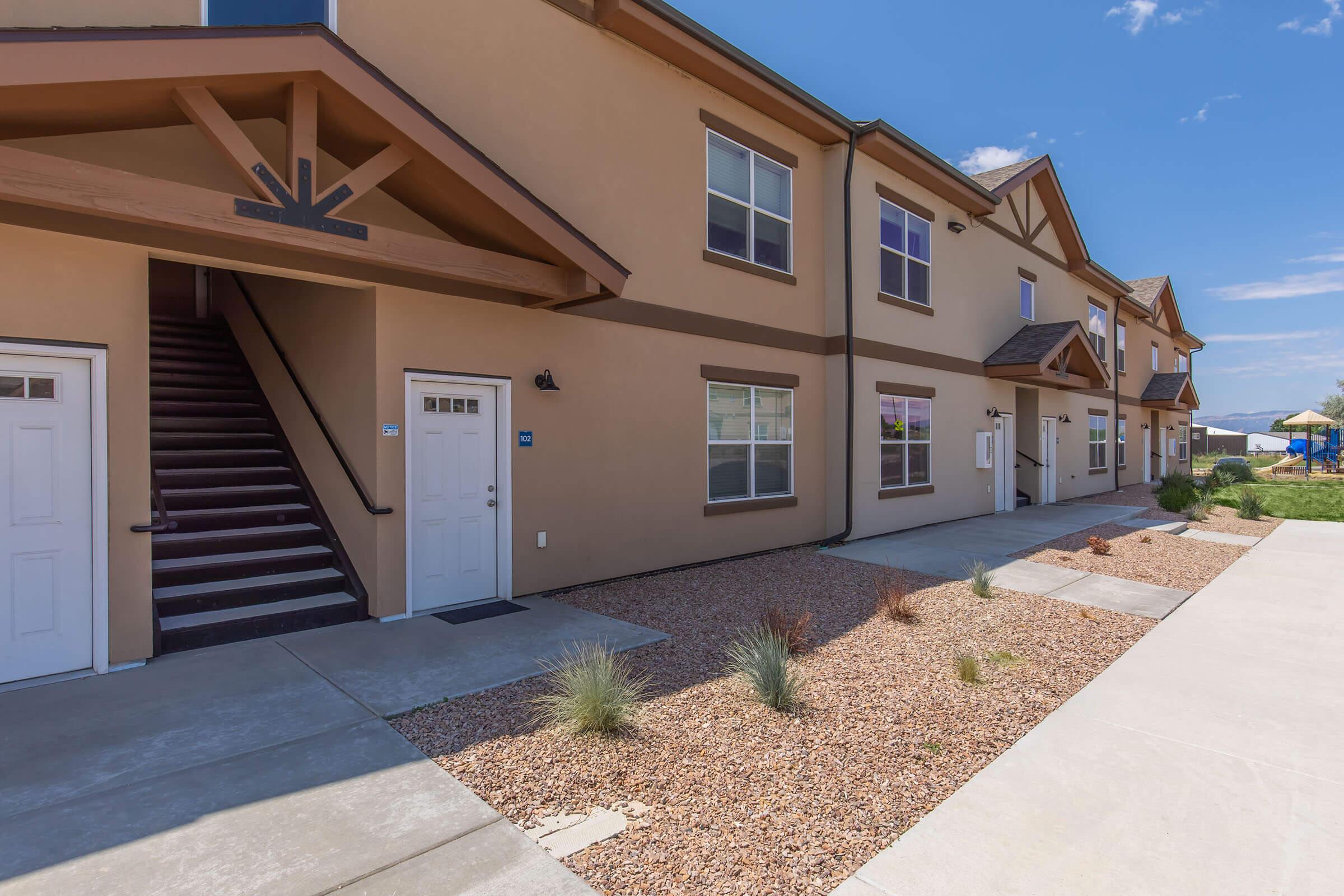Two-story building exterior with a stucco finish and wooden accents. There are two entrances visible, with an exterior staircase leading to the second floor. The landscaping includes desert plants and gravel. The sky is clear and blue, suggesting a sunny day.