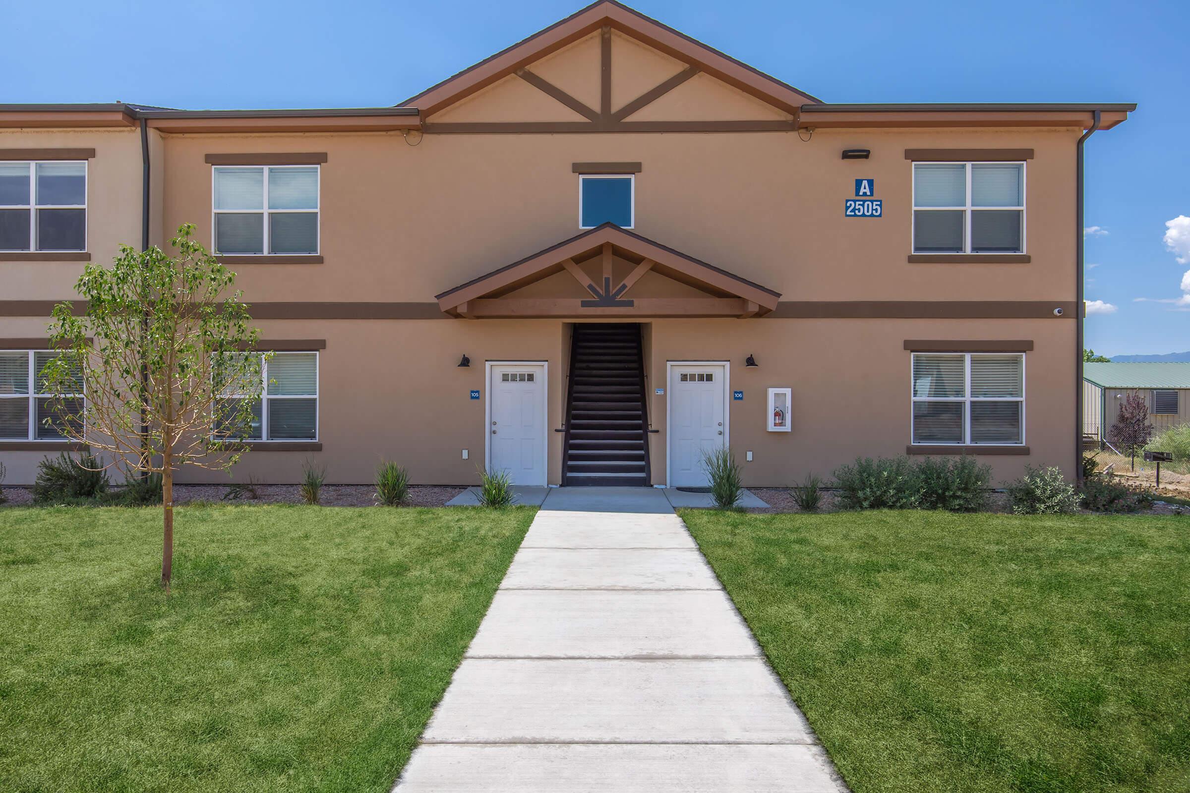 A modern two-story apartment building featuring a walkway leading to two entrances, surrounded by grass and small bushes. The building has a light brown exterior with a decorative gable and large windows. There is a visible address sign above the door and a clear blue sky in the background.