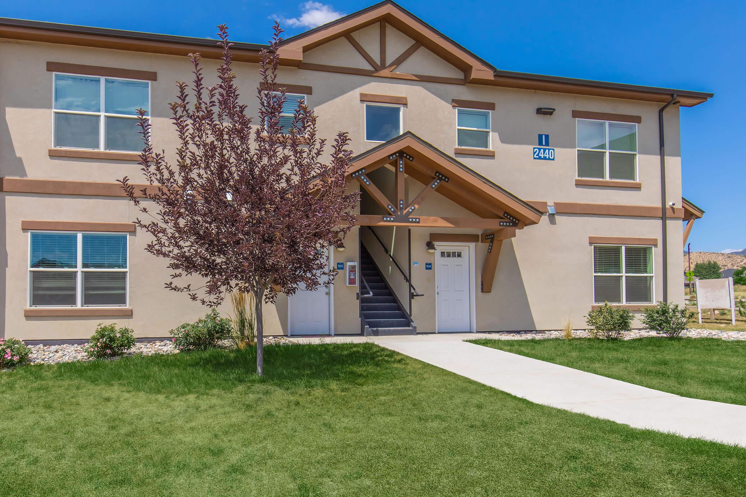 A two-story residential building with a light-colored exterior and brown trim. The building features a covered entrance with stairs leading up to the doors. A small flowering tree is in front, and the lawn is well-maintained with green grass. Clear blue skies are in the background.