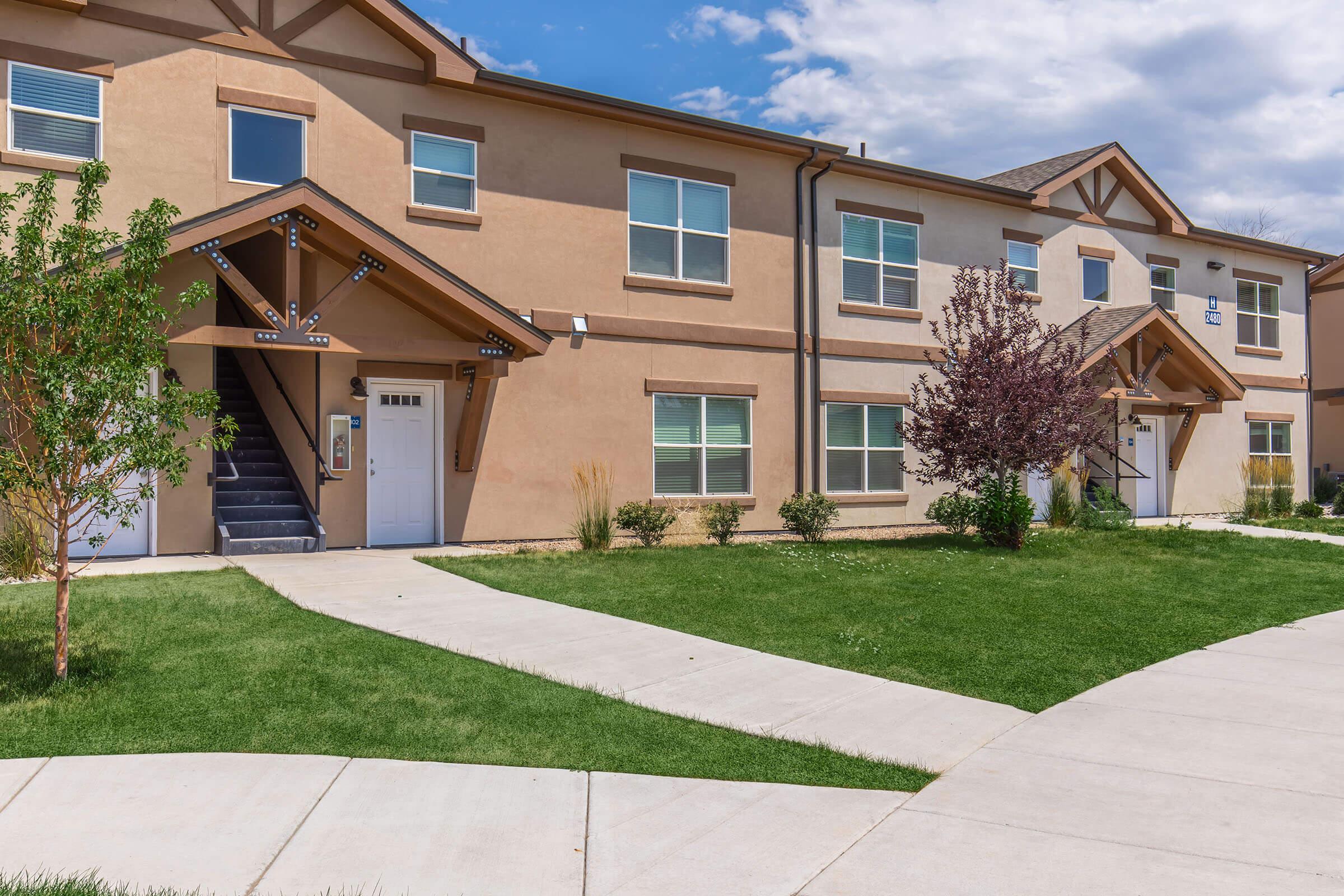 A modern two-story residential building with tan exterior walls and white trim. The entrance features a covered porch and stairs leading to the upper level. Lush green grass and decorative plantings line the walkway, creating a welcoming atmosphere. A clear blue sky with scattered clouds is visible above.