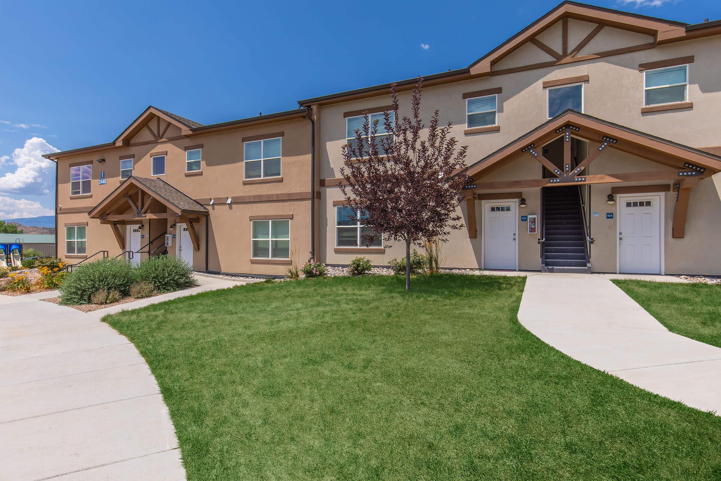 A two-story residential building with a modern design, featuring beige exterior walls, three entrance doors, and landscaped greenery in the foreground. A staircase leads to the upper level, and there are bushes and flowers around the property, under a blue sky with a few clouds.
