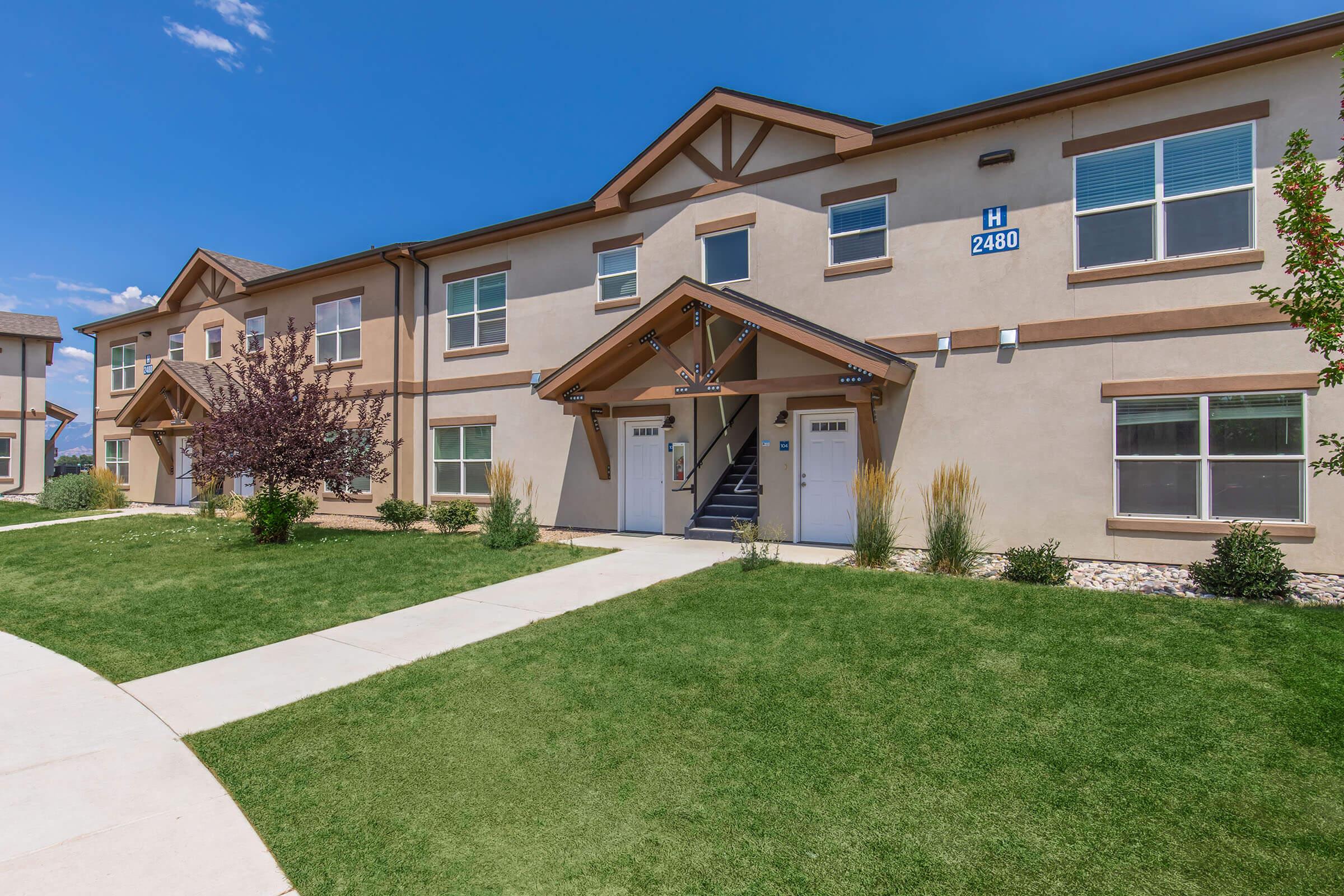 A modern two-story residential building with a welcoming entrance, featuring two front doors, well-maintained landscaping, and a clear blue sky. The facade has a beige color with decorative wooden accents, and the surrounding area includes green grass and small shrubs.