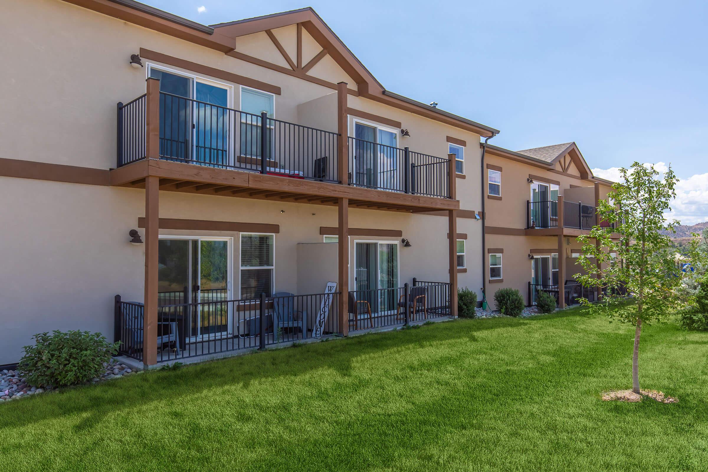 Two-story residential building with balconies on the upper floor. The exterior features a light-colored facade with dark accents. A well-maintained grassy area with small shrubs and a young tree is in the foreground, providing a pleasant outdoor space. Clear blue sky and sunlight add to the inviting atmosphere.
