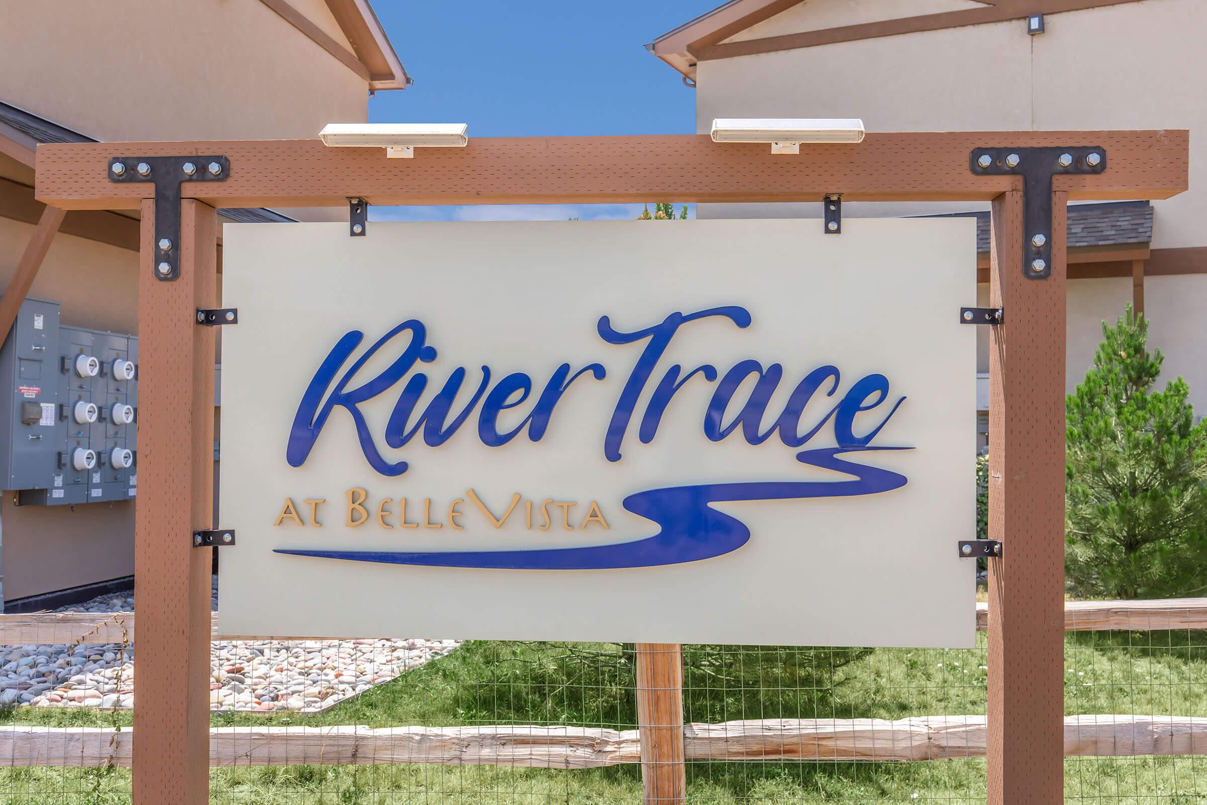 Sign for River Trace at Belle Vista, featuring stylized blue lettering, surrounded by a wooden frame. In the background, residential buildings and utility meters are visible. The area appears landscaped with decorative stones and greenery.