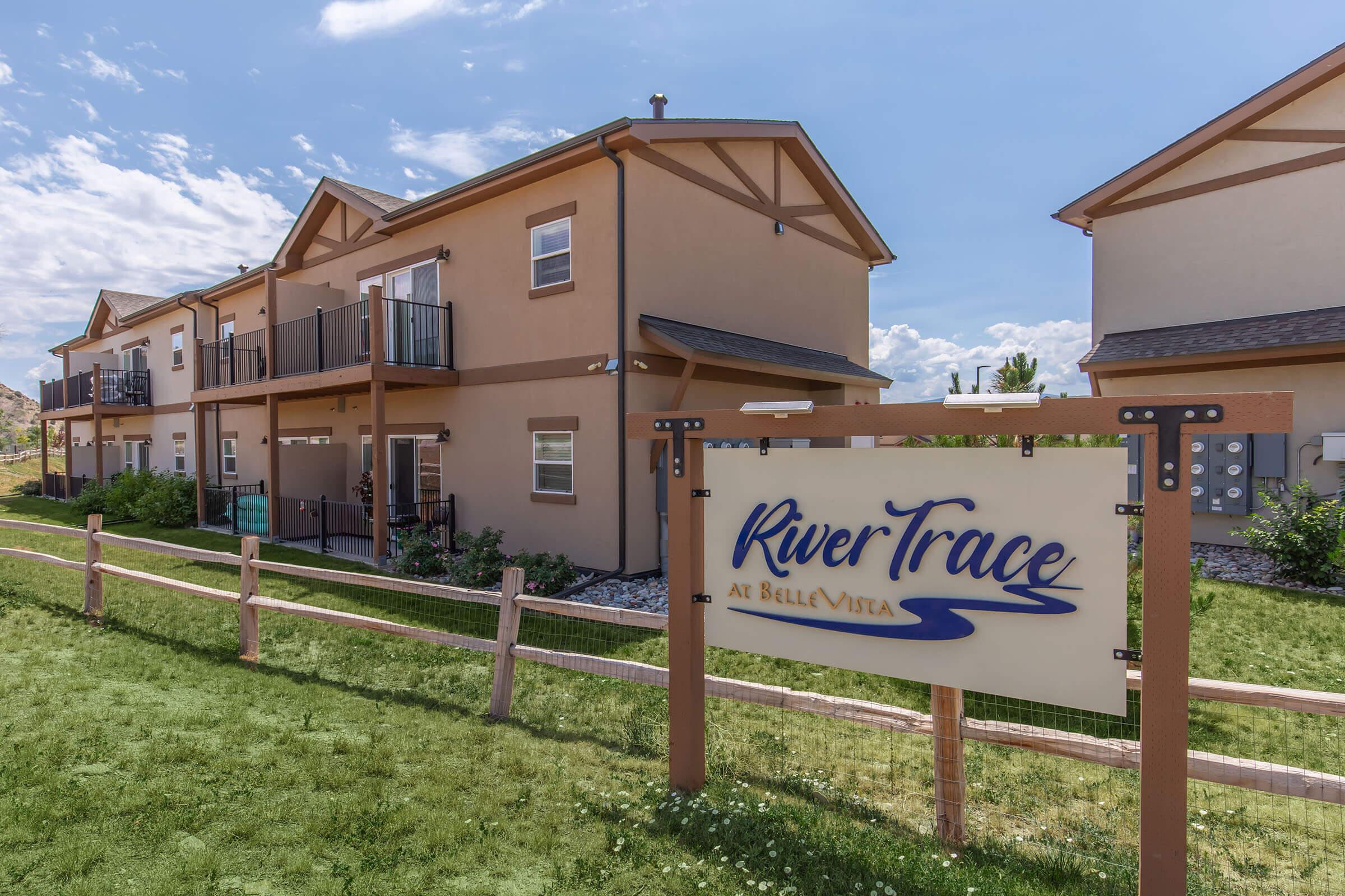 A view of the RiverTrace apartment complex at BelleVista, featuring multiple two-story buildings with balconies. A wooden fence surrounds a grassy area with a sign that reads "RiverTrace at BelleVista." The sky is partly cloudy, suggesting a pleasant day.