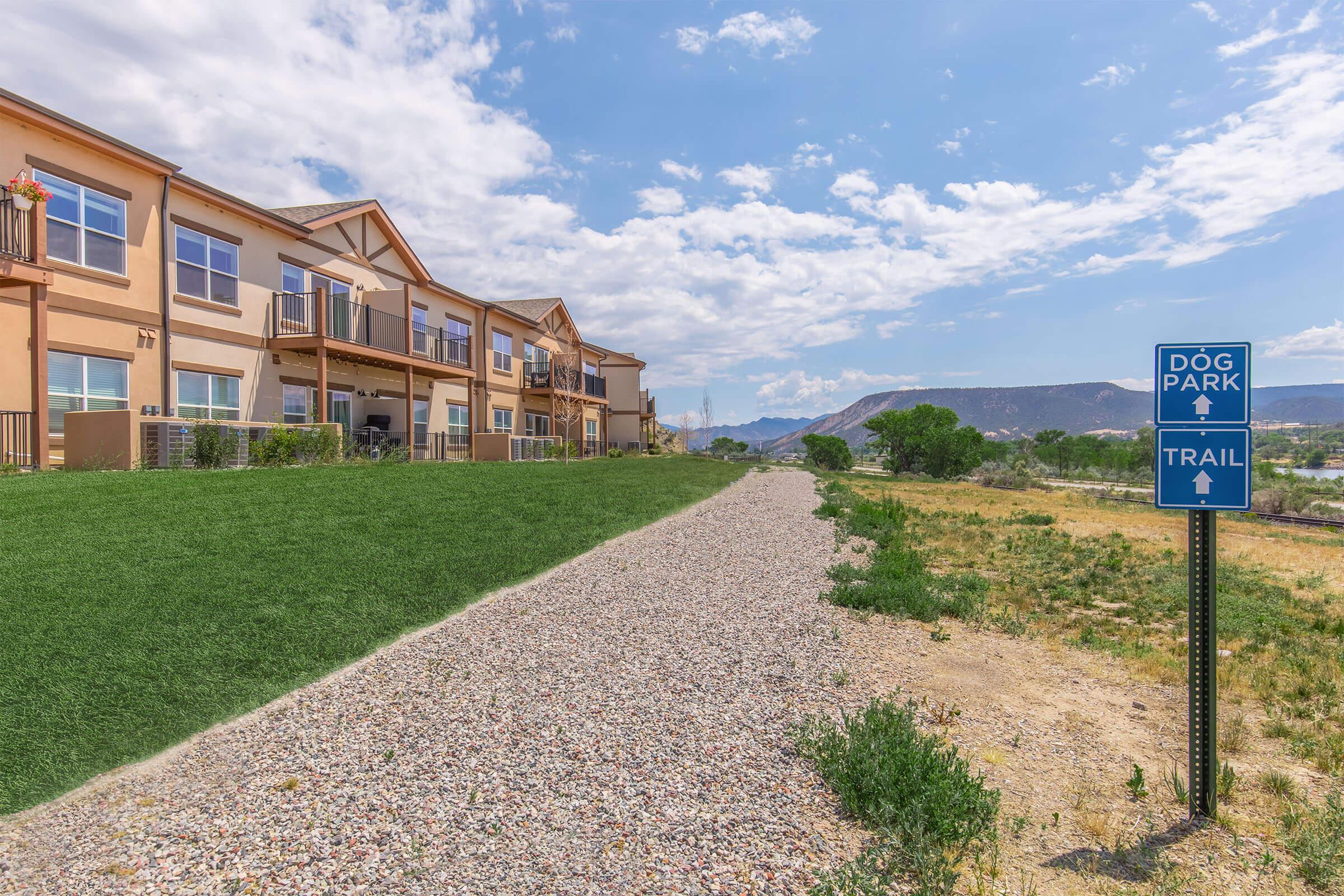 A scenic view of a residential area with multiple buildings on the left, lush green grass, and a gravel path leading to a sign for a dog park and trail. The sky is partly cloudy with distant mountains in the background, creating a peaceful outdoor atmosphere.