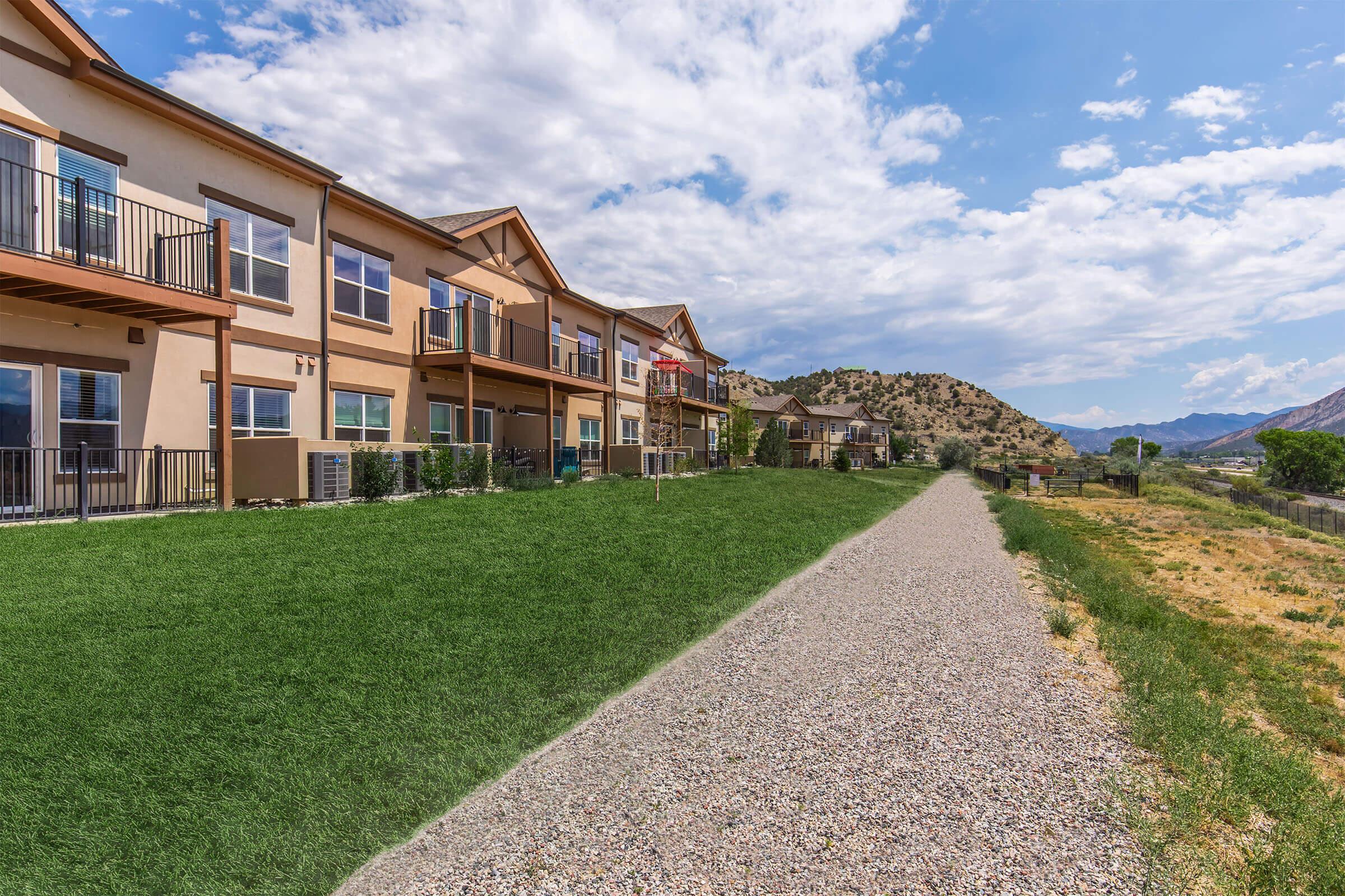 A scenic view of a residential building with a grassy area in the foreground and a gravel path leading towards mountains in the background. The sky is partly cloudy, adding depth to the landscape.