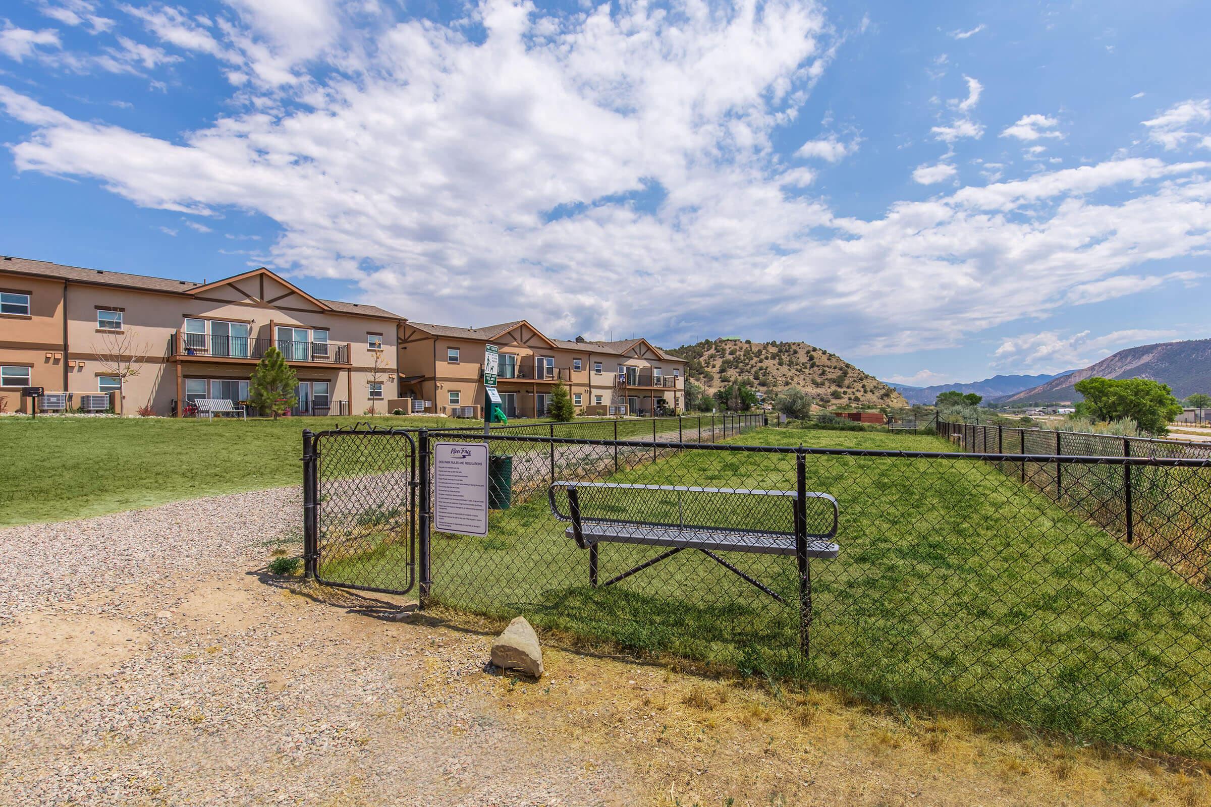 A well-maintained residential complex with several buildings in the background, surrounded by grassy areas and a fence. A sign is visible to the left, and there’s a bench near the entrance. The scene is set under a blue sky with scattered clouds and mountains in the distance.