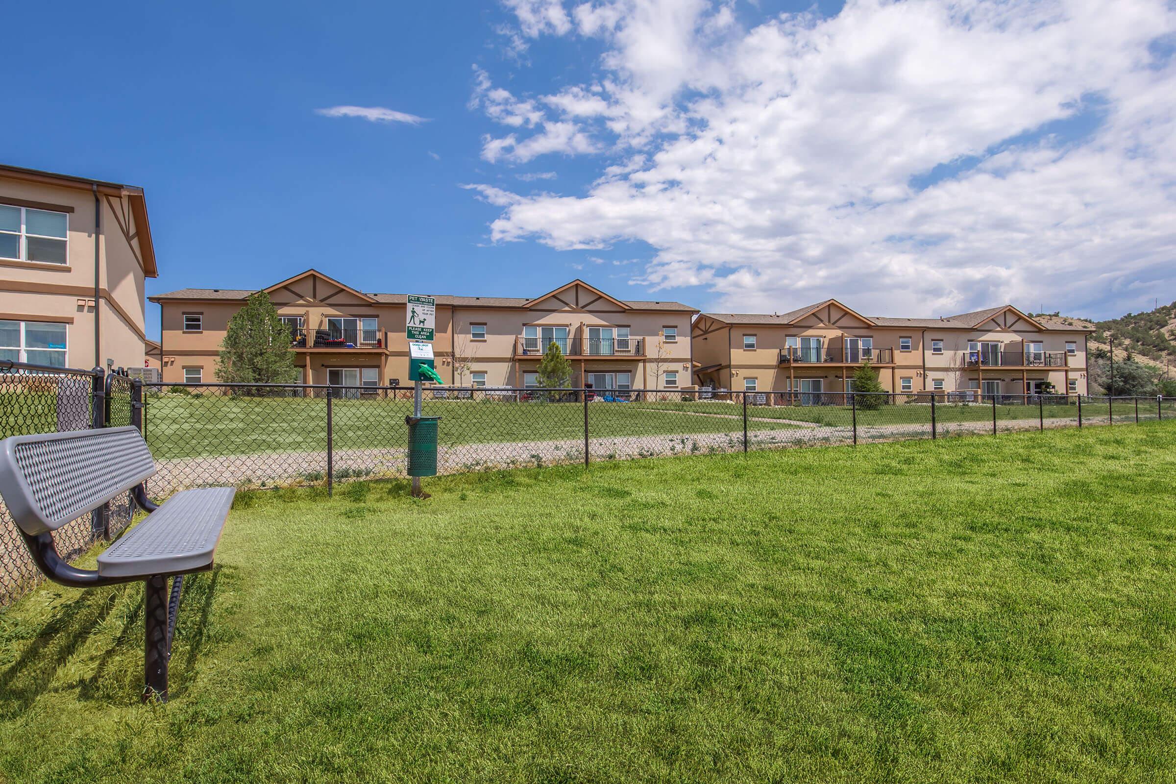 A view of an apartment complex from a grassy area, featuring well-maintained buildings with balconies. In the foreground, there is a gray bench beside a fenced area, and a clear blue sky with some clouds overhead. The landscape is inviting with green lawns and a tidy environment.