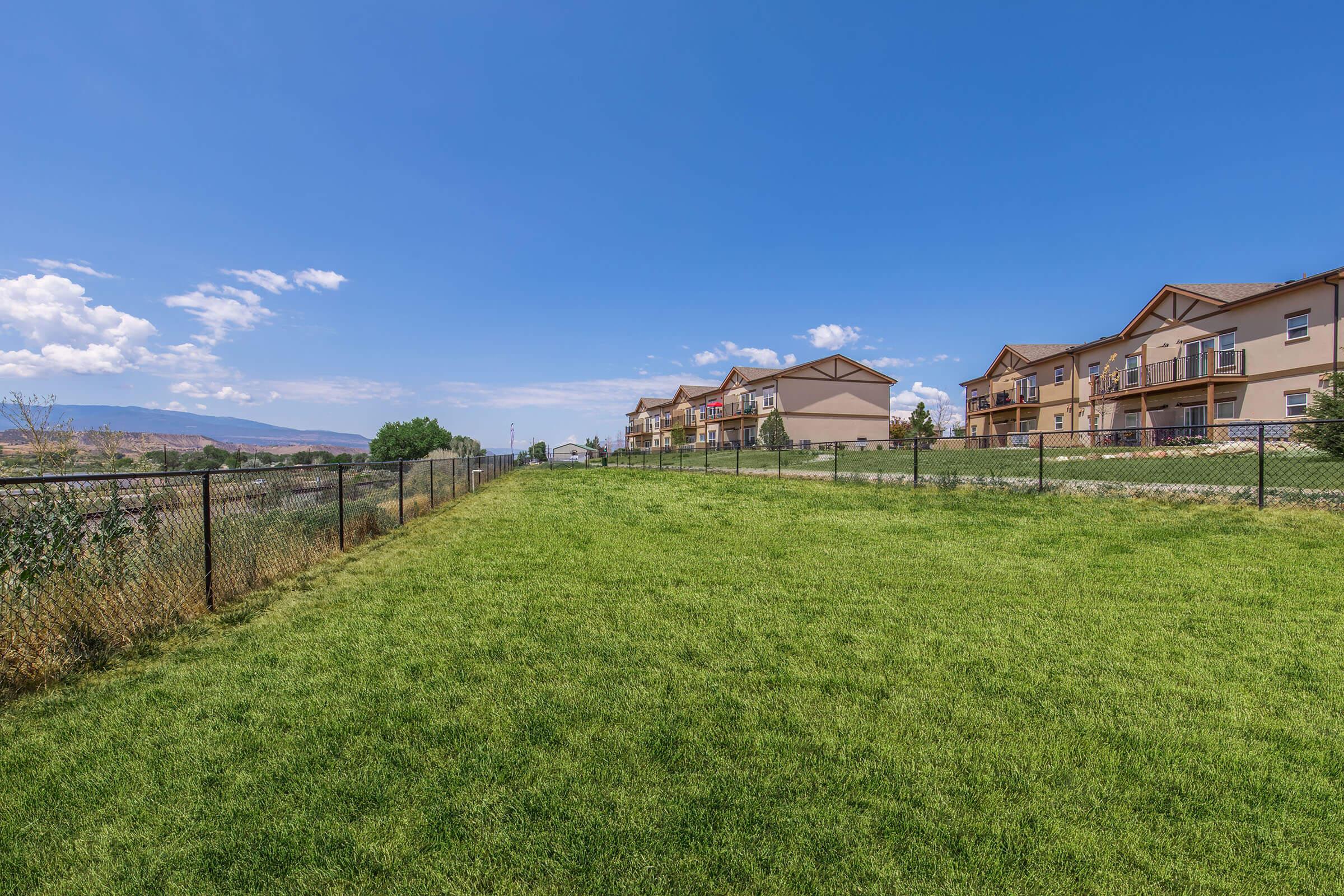 A spacious green lawn bordered by a black fence, with apartment buildings in the background under a bright blue sky. Fluffy white clouds are visible. The scene conveys a peaceful residential area with ample outdoor space.