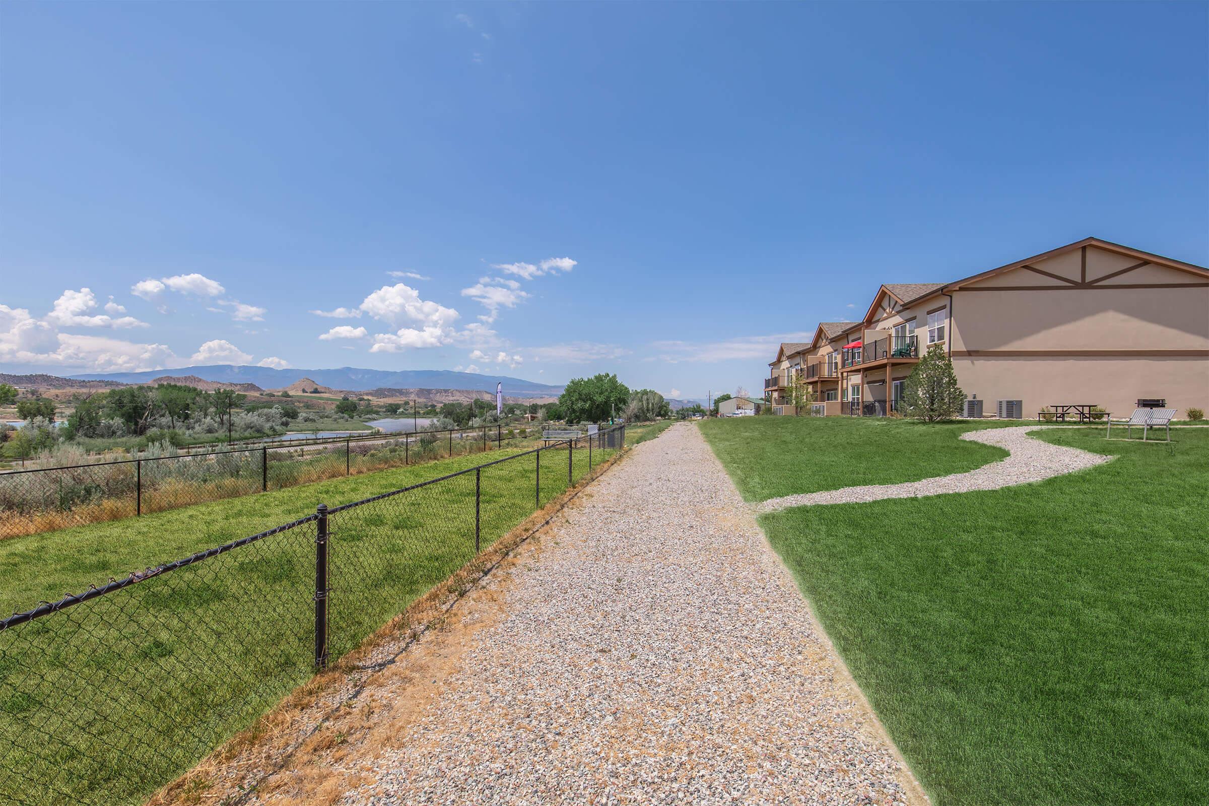 A scenic view of a gravel pathway lined with a black fence, leading through green grass and toward a row of multi-story buildings. In the background, rolling hills and a blue sky with scattered clouds are visible. The landscape has a serene, natural essence.