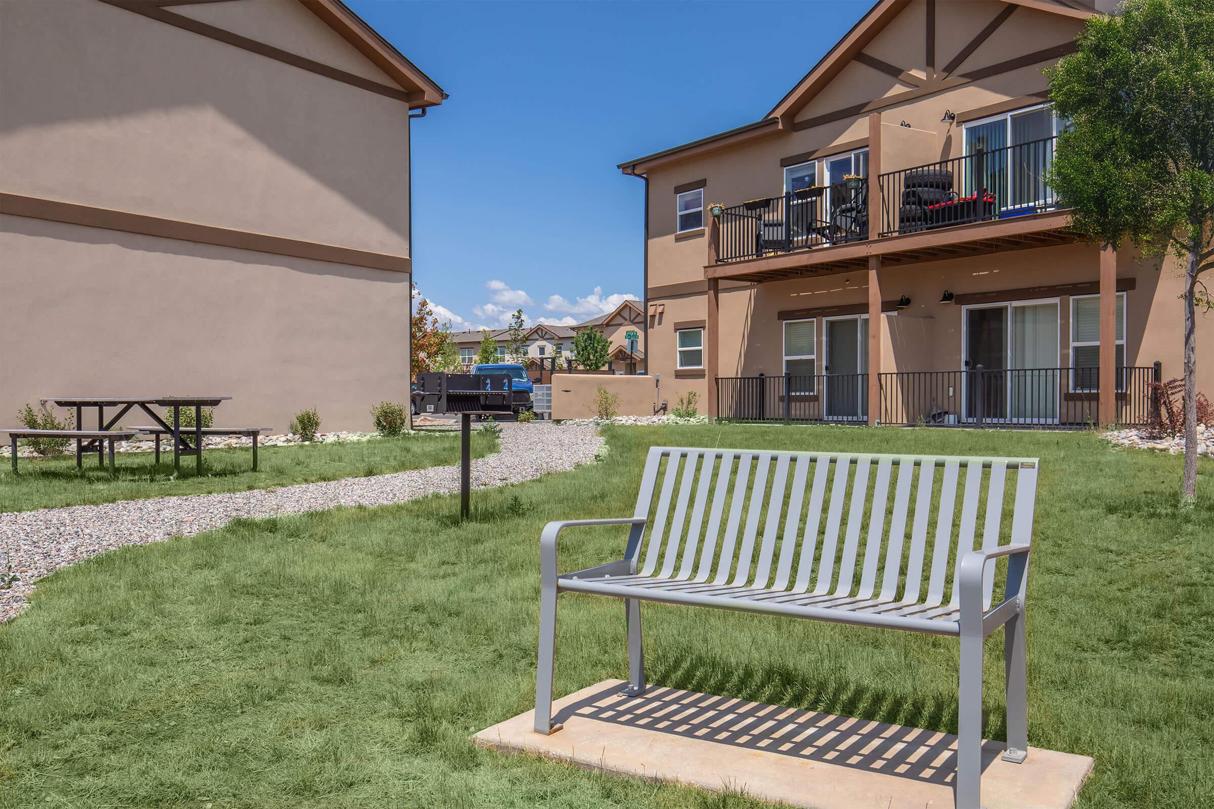 A silver metal bench sits on a grassy area in front of two modern multi-unit residential buildings. The scene includes a gravel path, a picnic table in the background, and clear blue skies with a few clouds, creating a serene outdoor space.