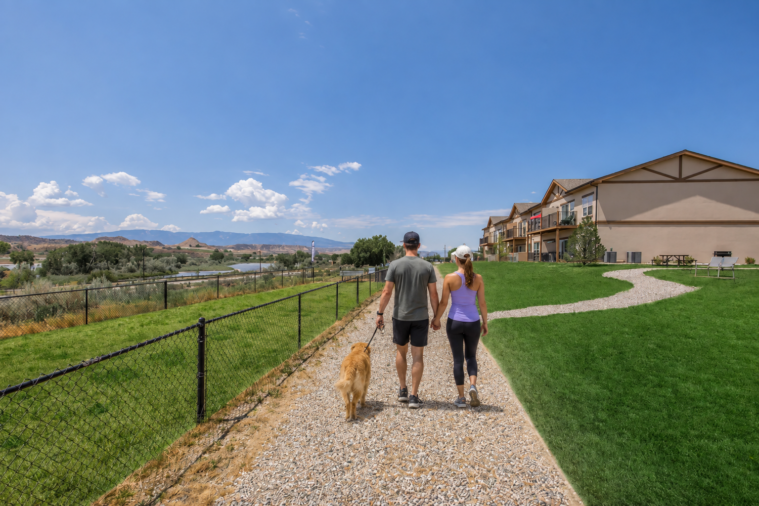 A man and a woman walking a golden retriever along a gravel path in a scenic area. They are surrounded by green grass and a fence, with colorful buildings and mountains in the background under a bright blue sky with clouds. The atmosphere is relaxed and sunny.