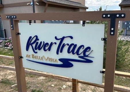 Sign for "River Trace at BelleVista" featuring elegant blue lettering on a white background, mounted on a wooden frame. The sign is set against a backdrop of landscaping and buildings, indicating a residential community.