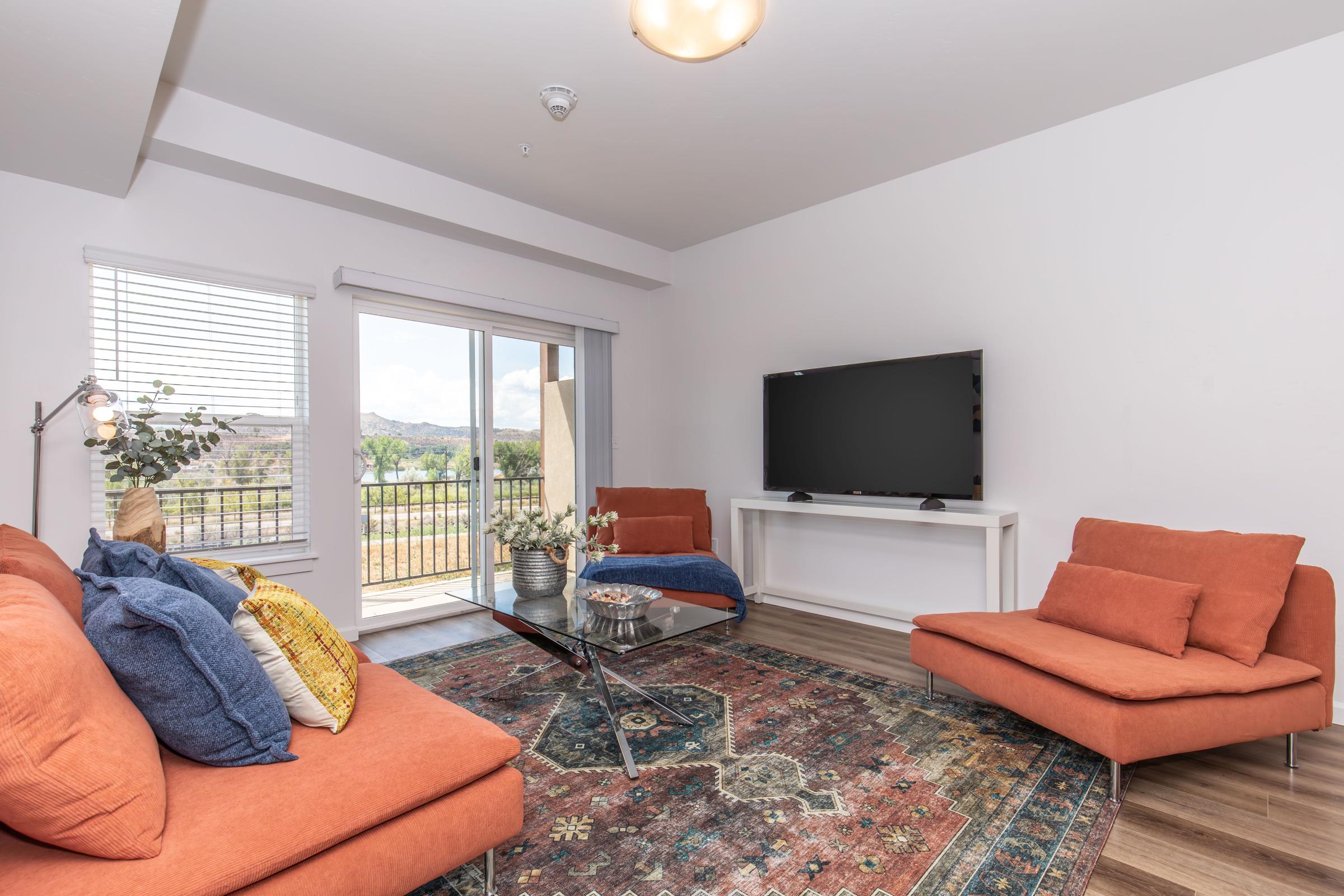 A cozy living room featuring two orange sofas, a coffee table with decorative items, and a large flat-screen TV on a white stand. The room has large windows with blinds and offers a view of outdoor greenery. A colorful area rug enhances the inviting atmosphere.