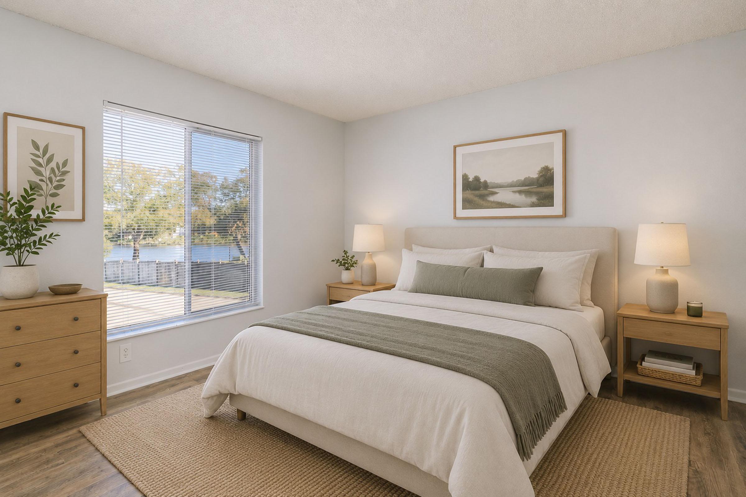 A serene bedroom featuring a soft bed with light bedding and a green throw. Two wooden nightstands with lamps are on either side, and a dresser is against the wall. A large window offers a view of trees and water outside, while a neutral color palette creates a calming atmosphere.