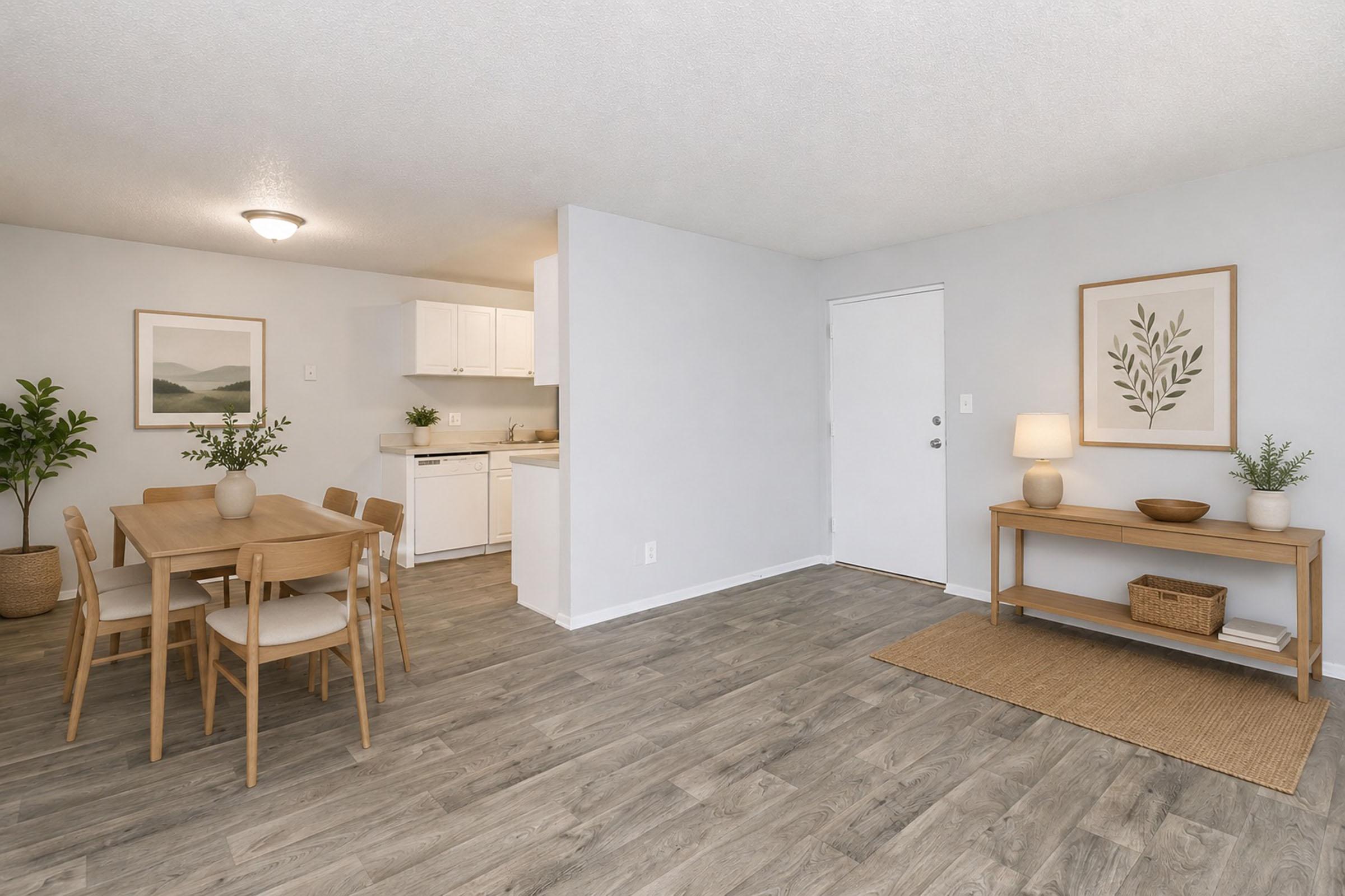 A bright and modern open-plan living space featuring a dining area with a wooden table and chairs, a small kitchen with white cabinetry, and a welcoming entryway. Decorative plants and minimalistic decor add a cozy touch to the neutral color scheme and wooden flooring.