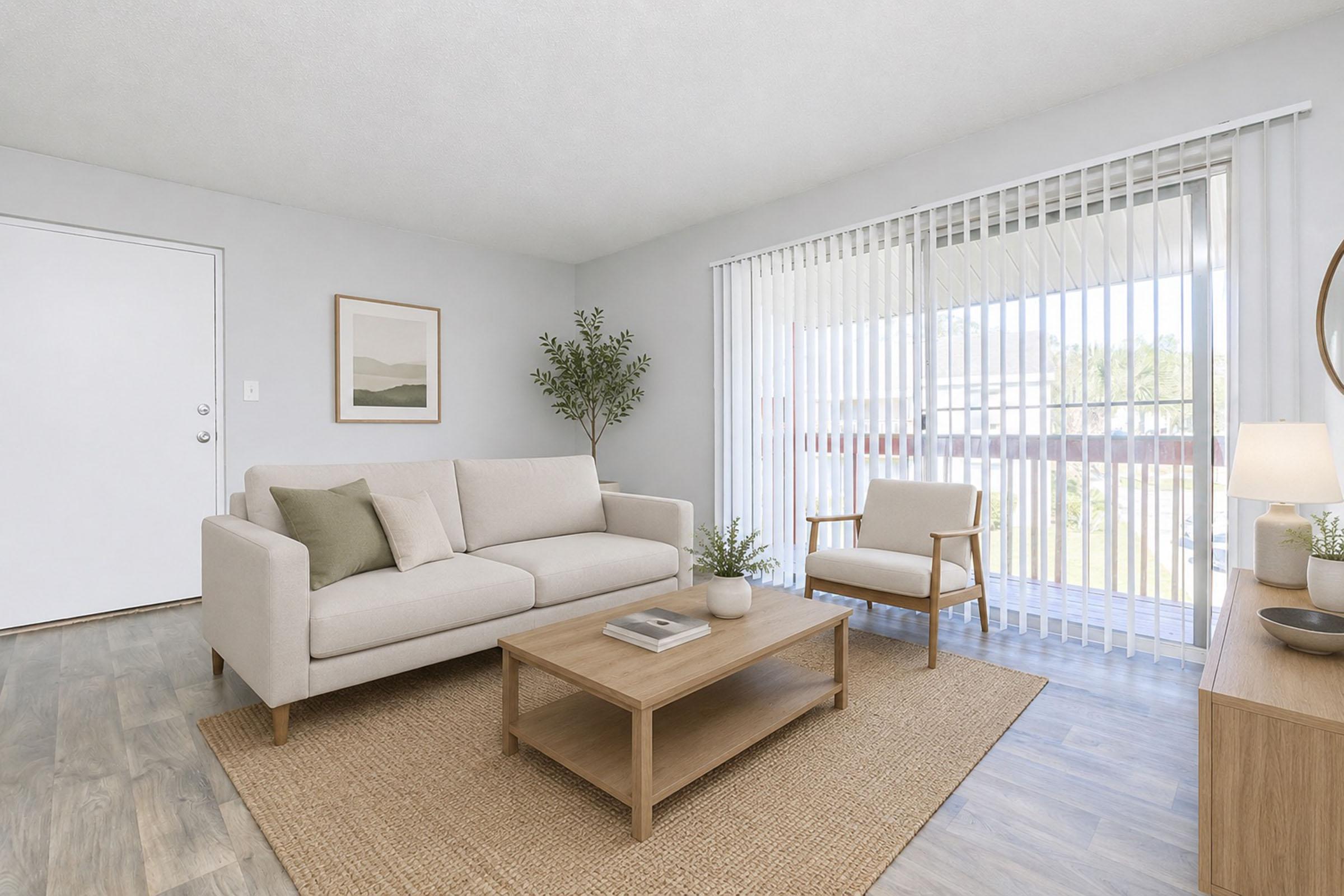 A cozy, modern living room featuring a light-colored sofa, a wooden coffee table, and an armchair. Natural light streams in through large sliding glass doors adorned with vertical blinds. A simple wall art piece and a potted plant add a touch of greenery to the neutral decor.