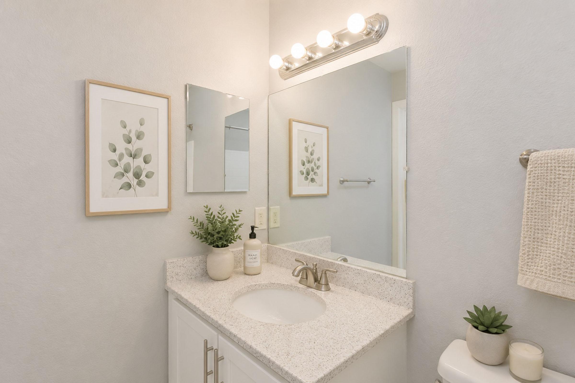 A modern bathroom with light gray walls, featuring a white vanity with a sink, a large mirror, and decorative wall art. There is a plant on the counter and a towel hanging nearby. Soft lighting is provided by a fixture above the mirror, creating a bright and inviting atmosphere.