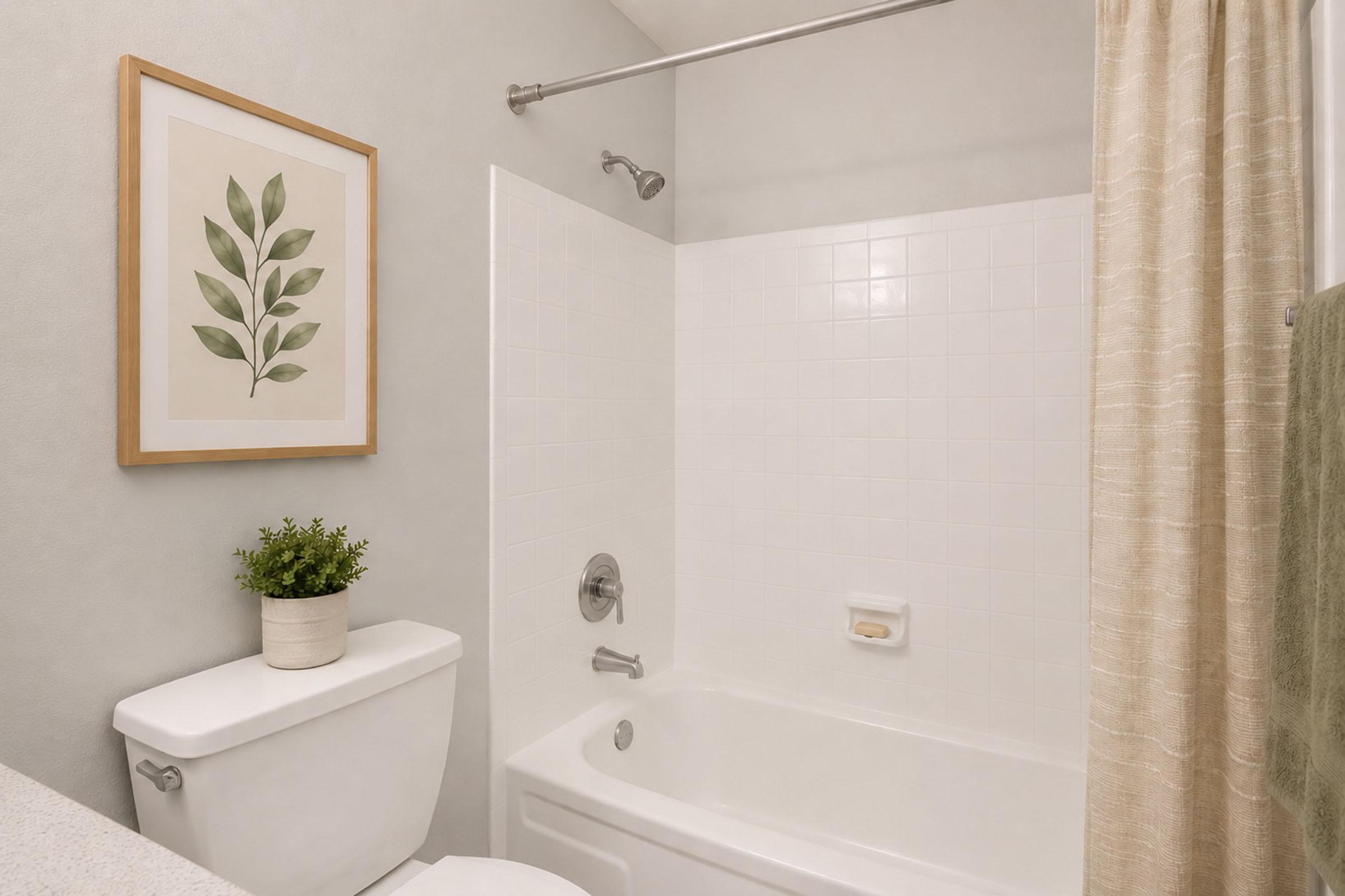 A clean and modern bathroom featuring a white bathtub with a shower, a light-colored shower curtain, a wall-mounted showerhead, a potted plant on the countertop, and a framed botanical print on the wall. The overall design is minimalistic and airy, with soft neutral tones.