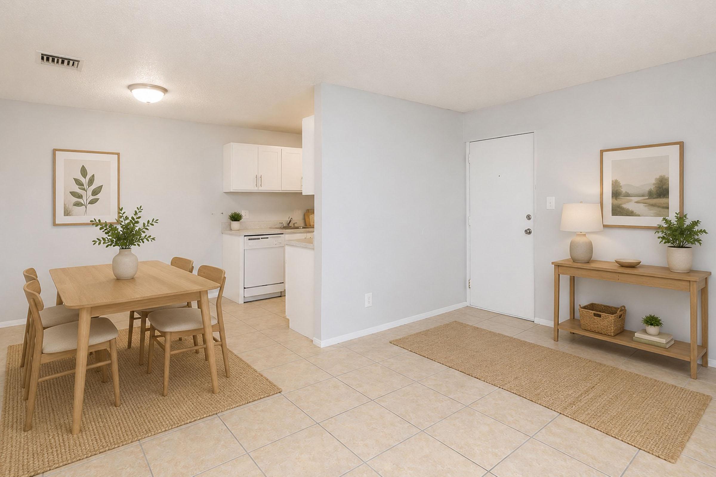 A bright and airy kitchen and dining area featuring a wooden dining table with four chairs, light-colored walls, and tiled flooring. The kitchen is partially visible with white cabinetry and appliances. Two framed botanical prints decorate the walls, and a console table with decorative items is positioned near the entrance.