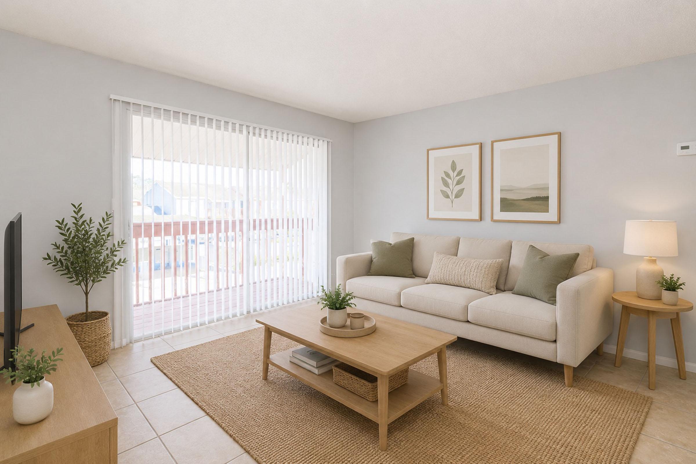 Bright and airy living room featuring a light-colored couch with decorative cushions, a coffee table, and a TV on a wooden stand. Natural light streams in through a sliding glass door with vertical blinds. Potted plants and framed botanical artwork add a touch of nature to the modern decor.