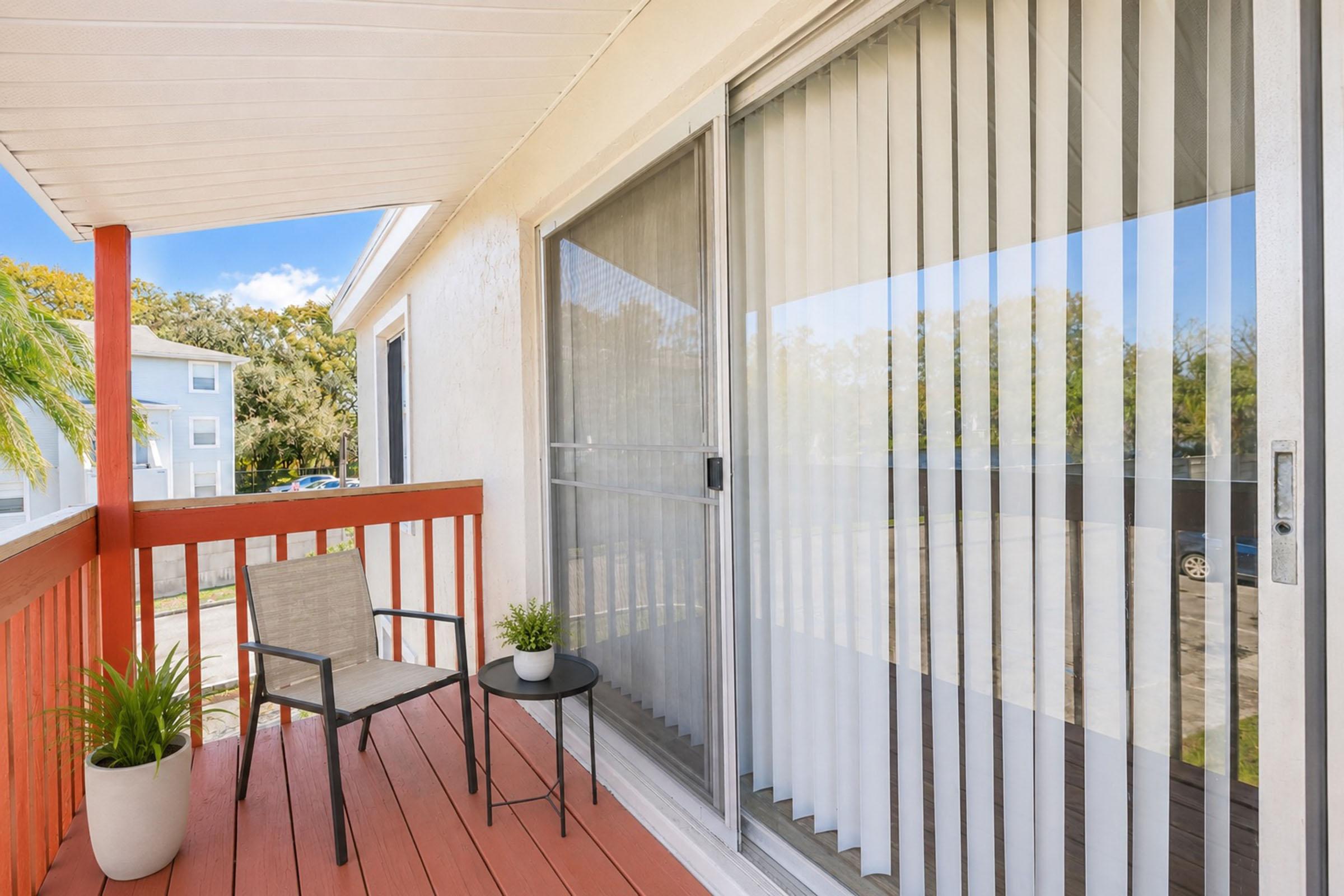 A small balcony with a red railing, featuring a chair and a round table. There are potted plants nearby, and large sliding glass doors with vertical blinds. In the background, there are hints of trees and buildings. The scene is bright and inviting, ideal for relaxation.