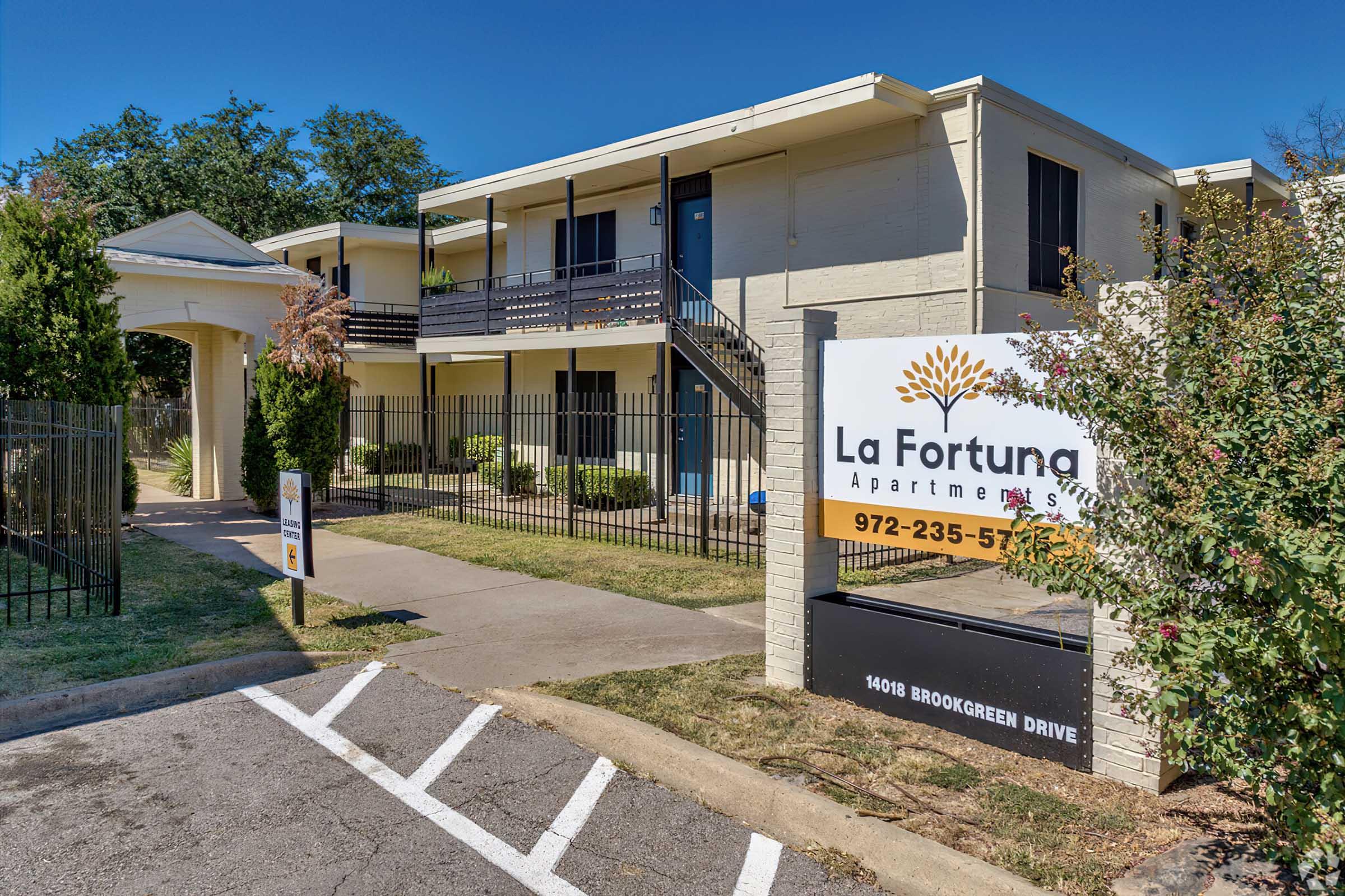 Exterior view of La Fortuna Apartments featuring a two-story building with a sign at the entrance. The sign displays the name, contact number, and address. Surrounding greenery includes trees and shrubs, with a paved walkway leading to the building entrance. Clear blue sky in the background.