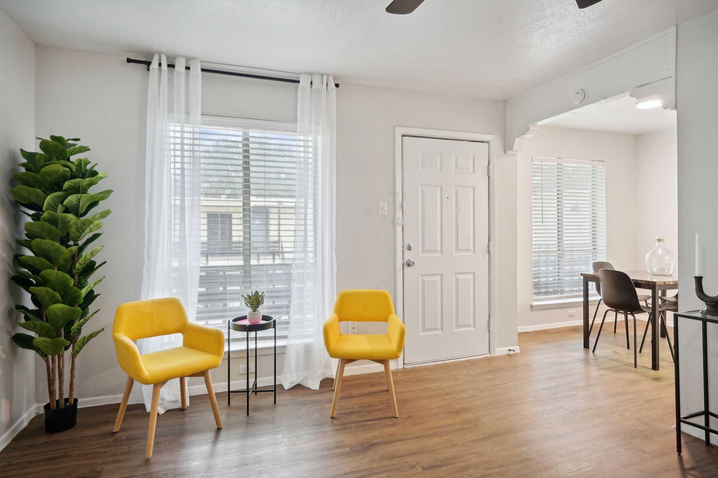 A bright, cozy room featuring two yellow chairs beside a small round table with a potted plant. Light streams in through large windows with sheer white curtains. A white door leads outside, and a dining area with a table and chairs is visible in the background, enhancing the inviting atmosphere.