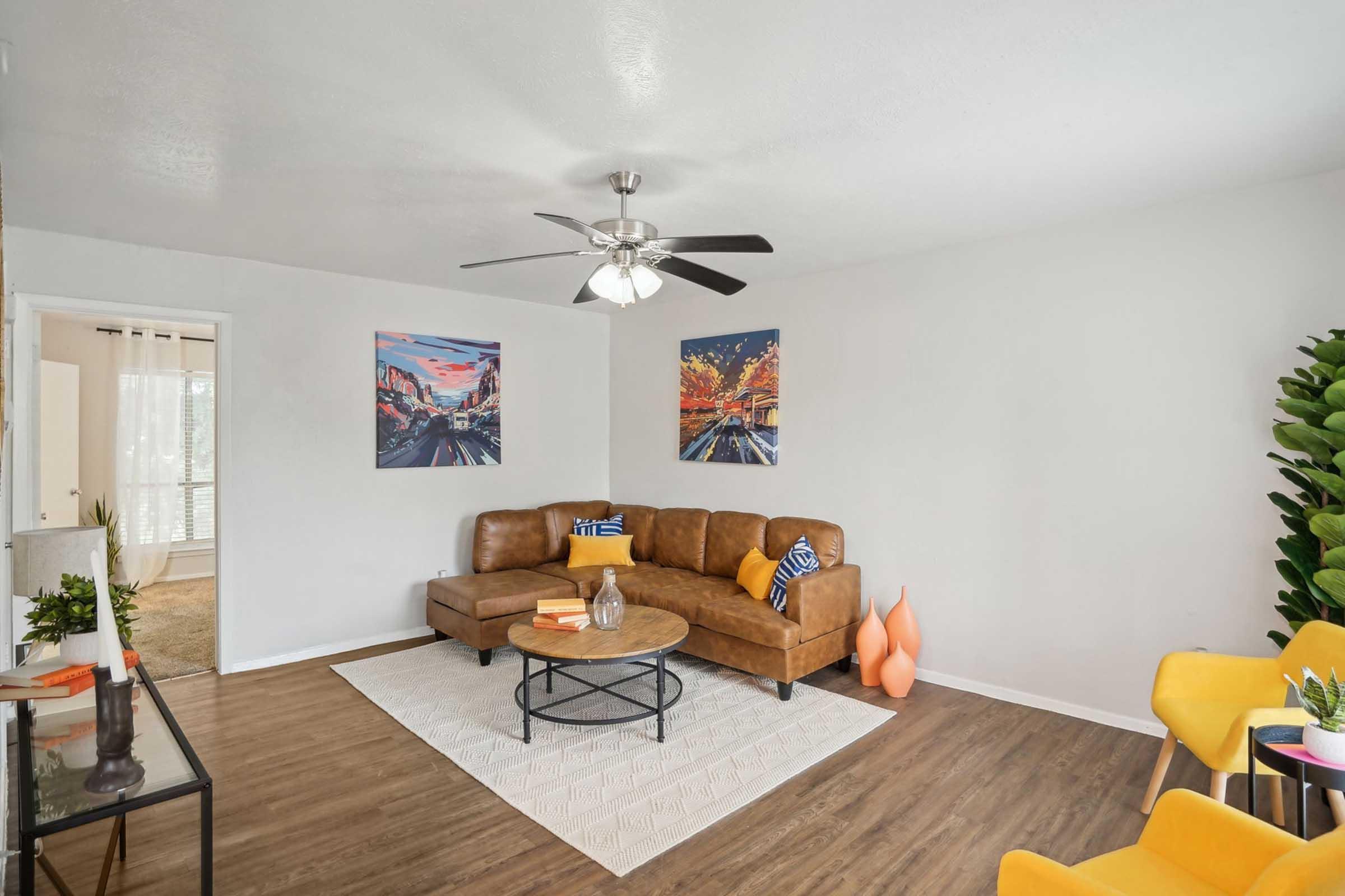 A cozy living room featuring a brown leather sectional couch with decorative pillows, a round coffee table, and vibrant artwork on the walls. There are orange vases, a potted plant, and yellow dining chairs, enhancing the inviting atmosphere. Soft natural light enters through a doorway, adding warmth to the space.