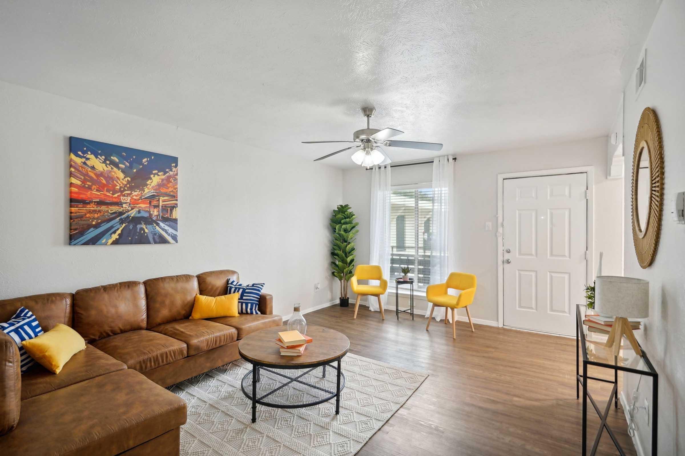 A cozy living room featuring a brown leather sectional sofa with colorful cushions, a round coffee table, and a patterned rug. A wall clock and framed artwork adorn the walls. There's a tall plant in the corner, two yellow accent chairs, and a door leading to a patio with large windows allowing natural light.