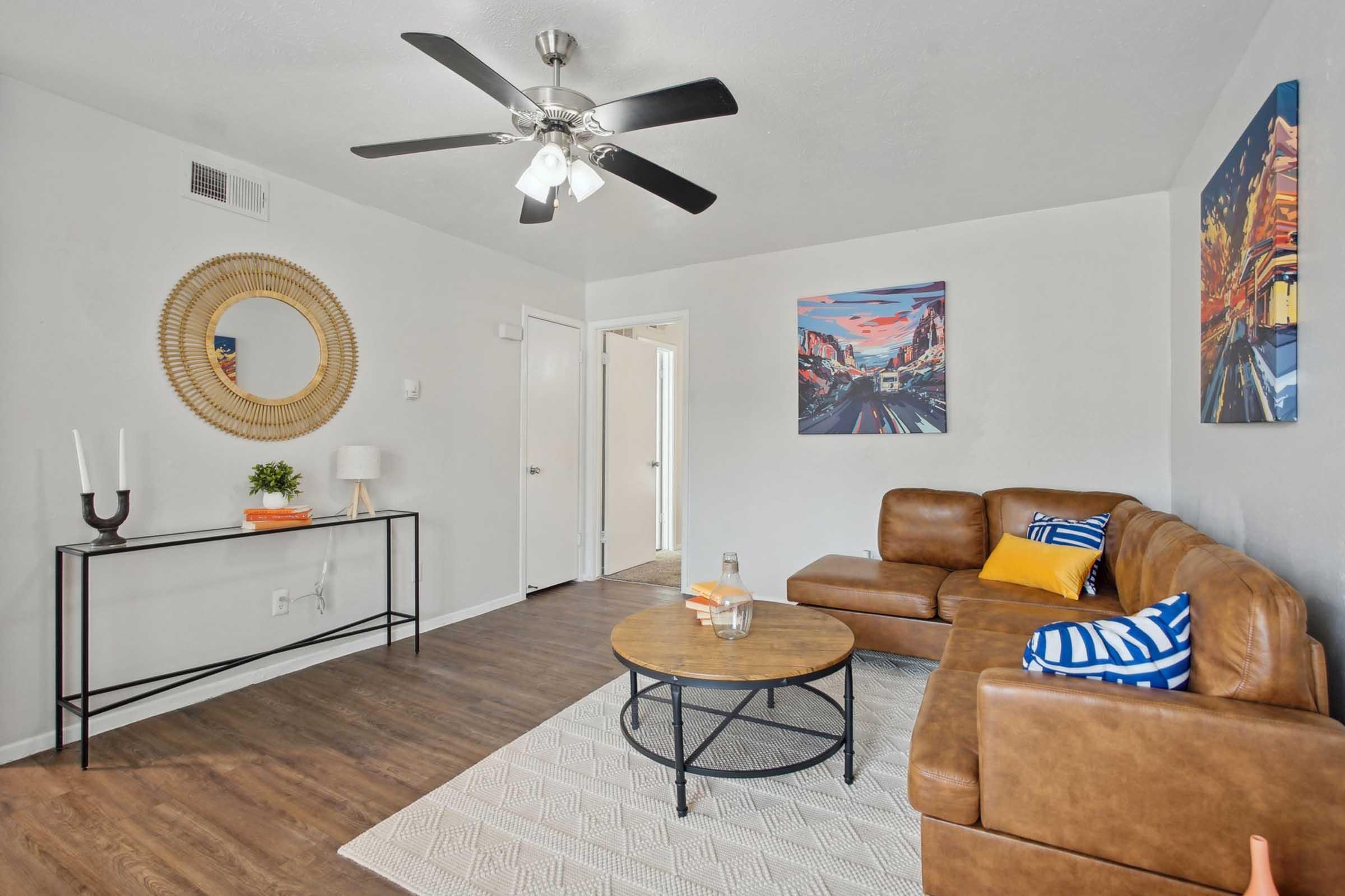 A cozy living room featuring a brown leather sectional sofa with blue and white striped cushions, a round coffee table, and a decorative area rug. The walls are painted light gray, adorned with two colorful art pieces. A ceiling fan and a small side table with a lamp complete the inviting space.