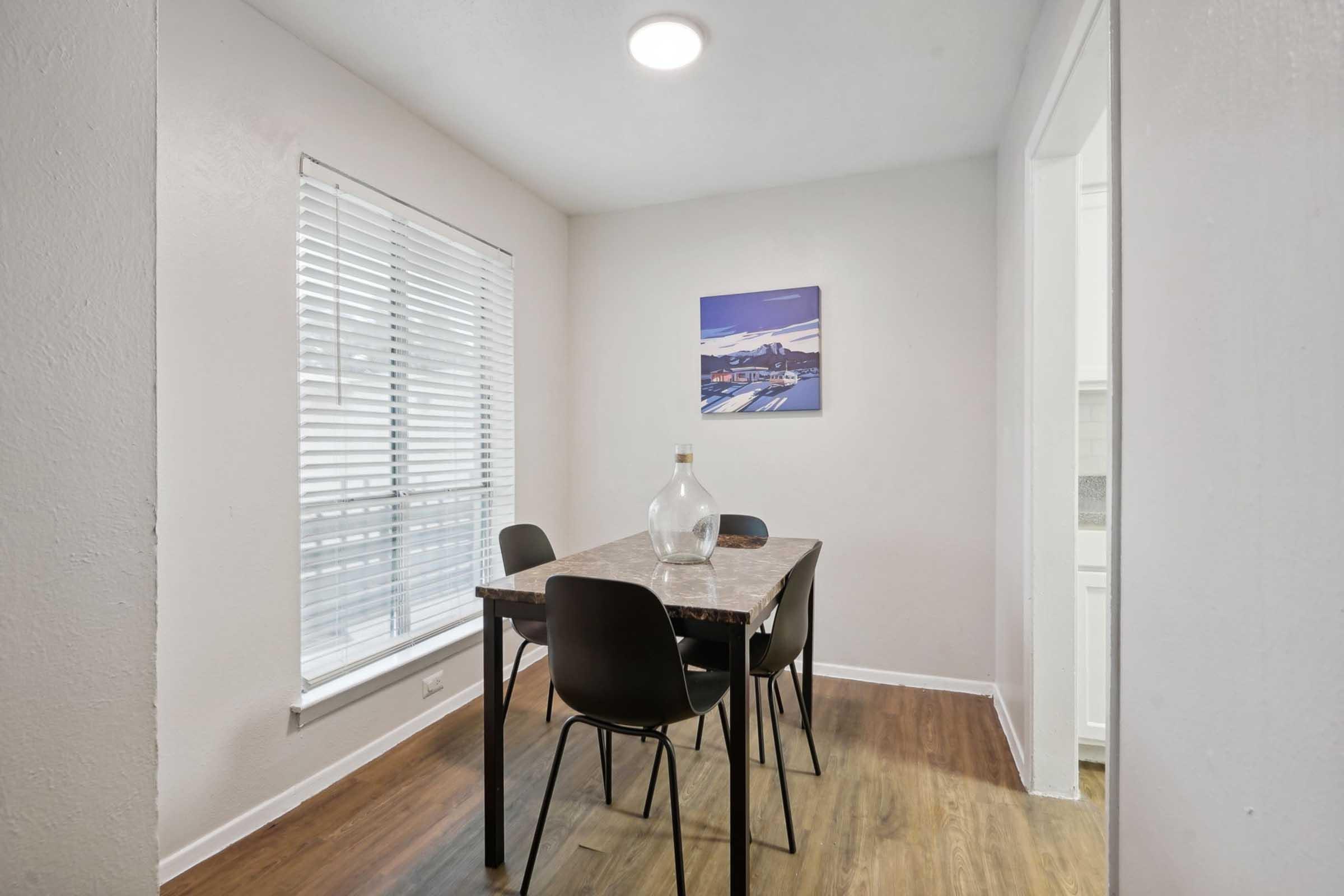 A bright dining area featuring a small table with four black chairs. A glass vase is on the table, and a modern artwork depicting a landscape hangs on the wall. Natural light streams in through a window with white blinds, enhancing the warm wooden floor and clean, minimalist decor.